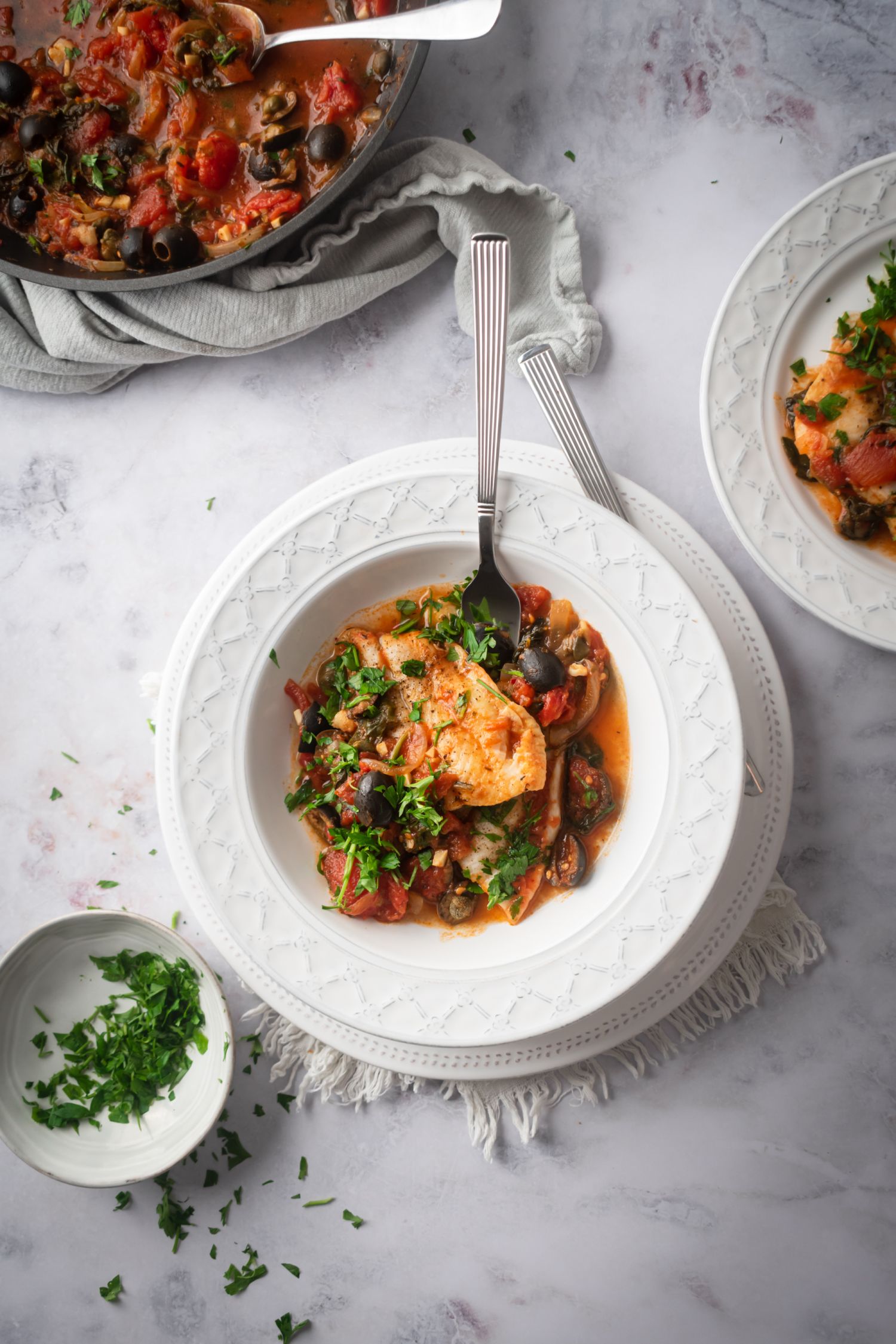 White fish with Mediterranean sauce in a white bowl with fresh parsley, olives, and capers.