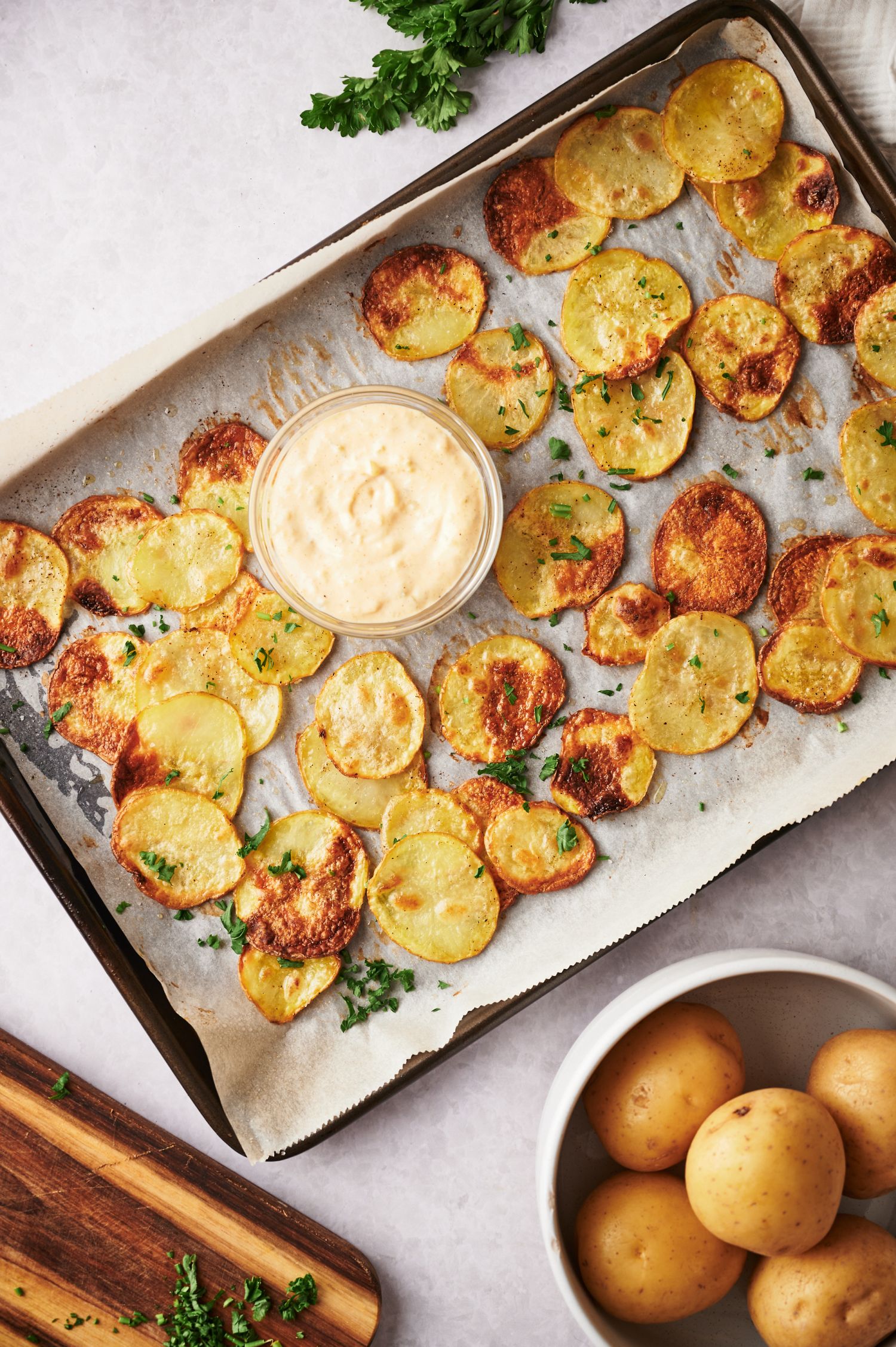 Golden brown potato chips on a baking sheet covered in parchment paper with herbs on the side.