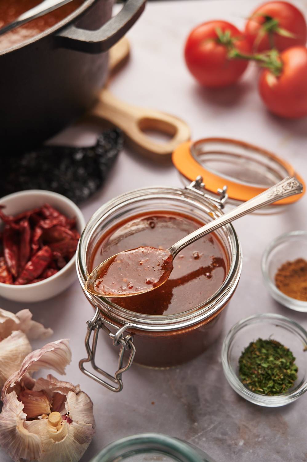 Homemade birria sauce in glass jar with dried chile peppers, garlic, and herbs on the side.