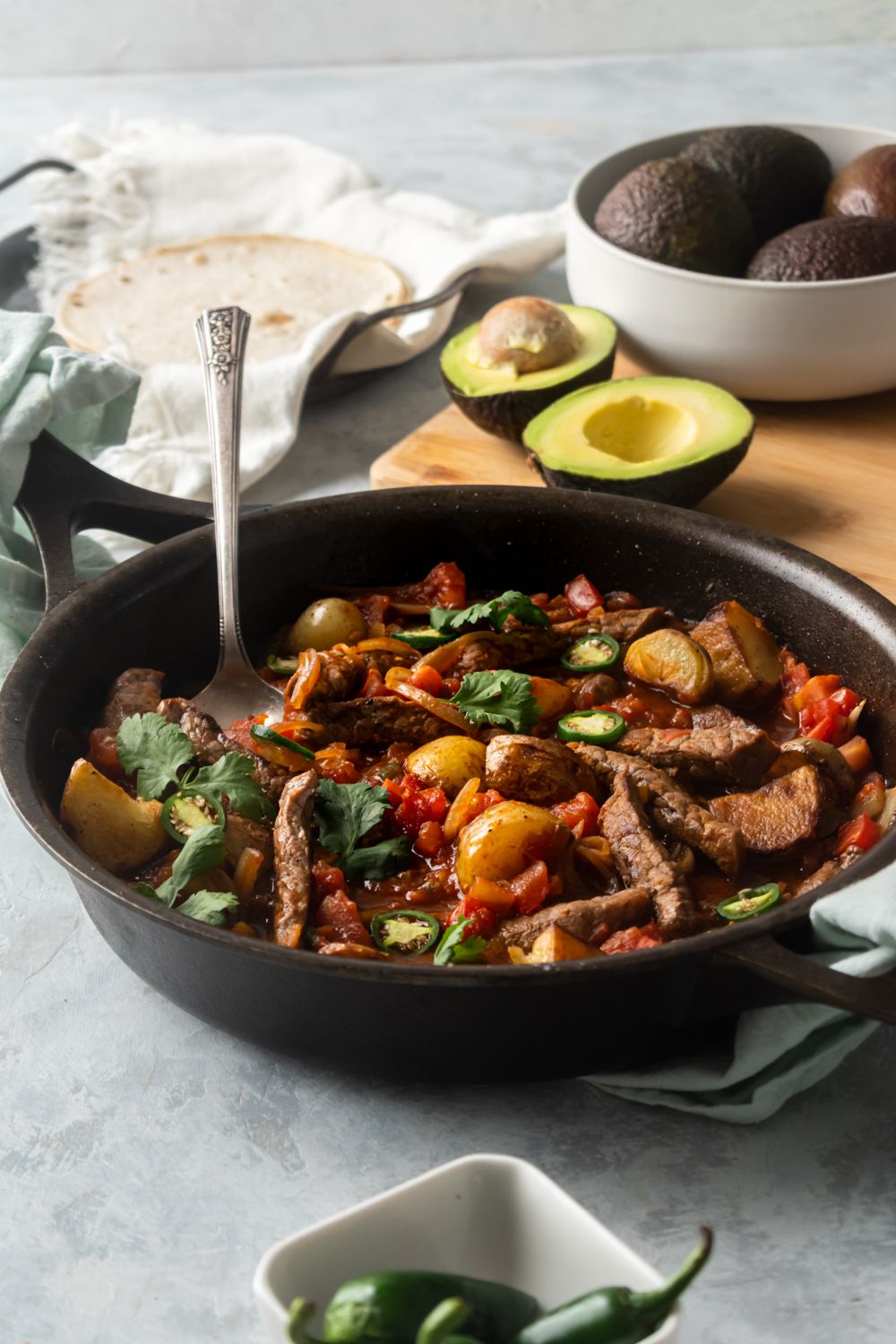 Mexican bistec ranchero served in a skillet with fresh avocados and tortillas on the side.