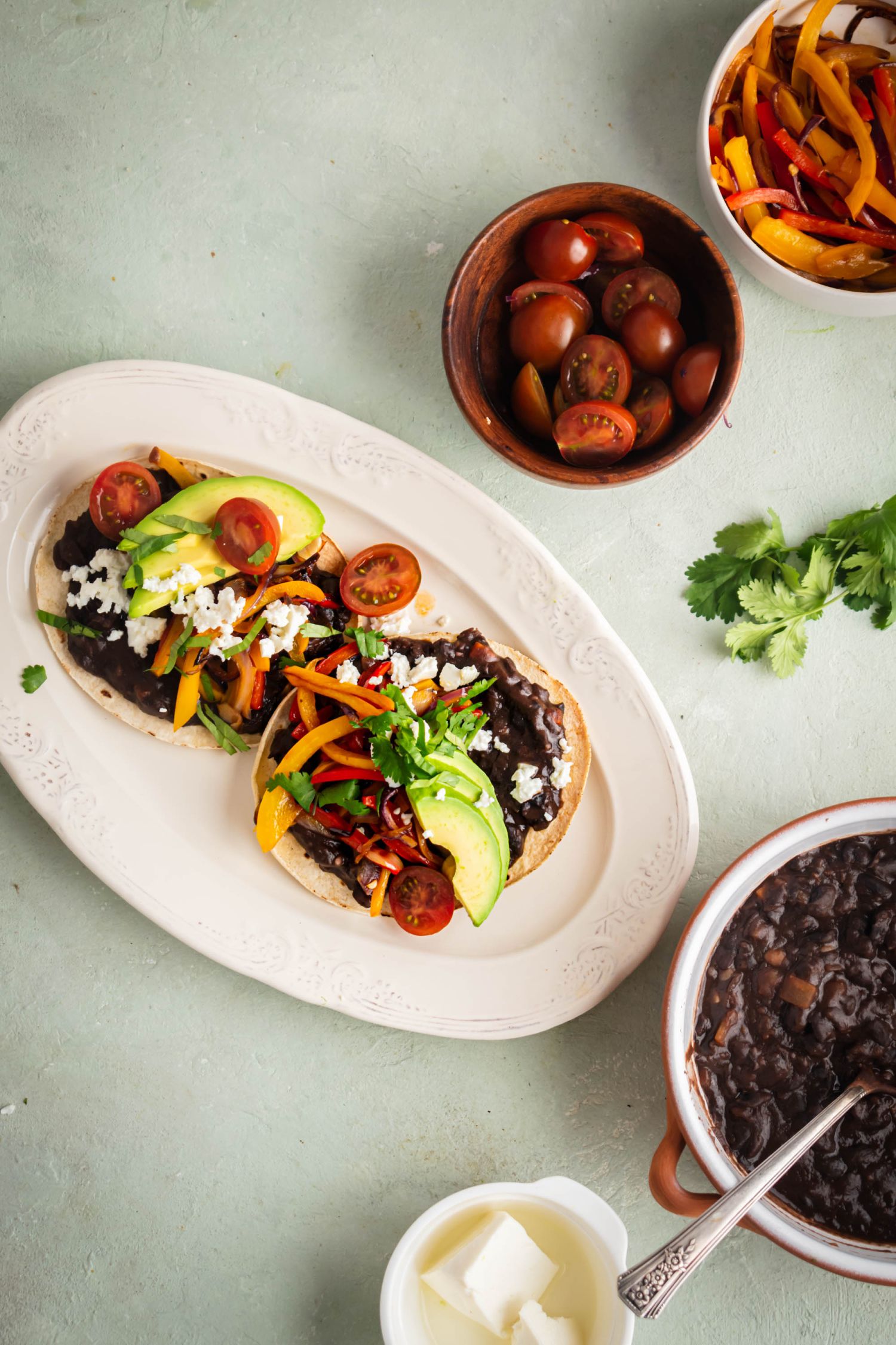 Homemade black bean tostadas on a white plate with tomatoes, peppers, red onions, avocado, cilantro, and cheese.