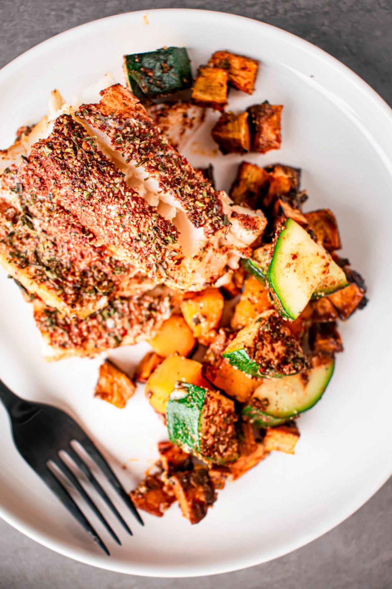 Blackened codfish with zucchini and sweet potatoes on a white plate with a fork.