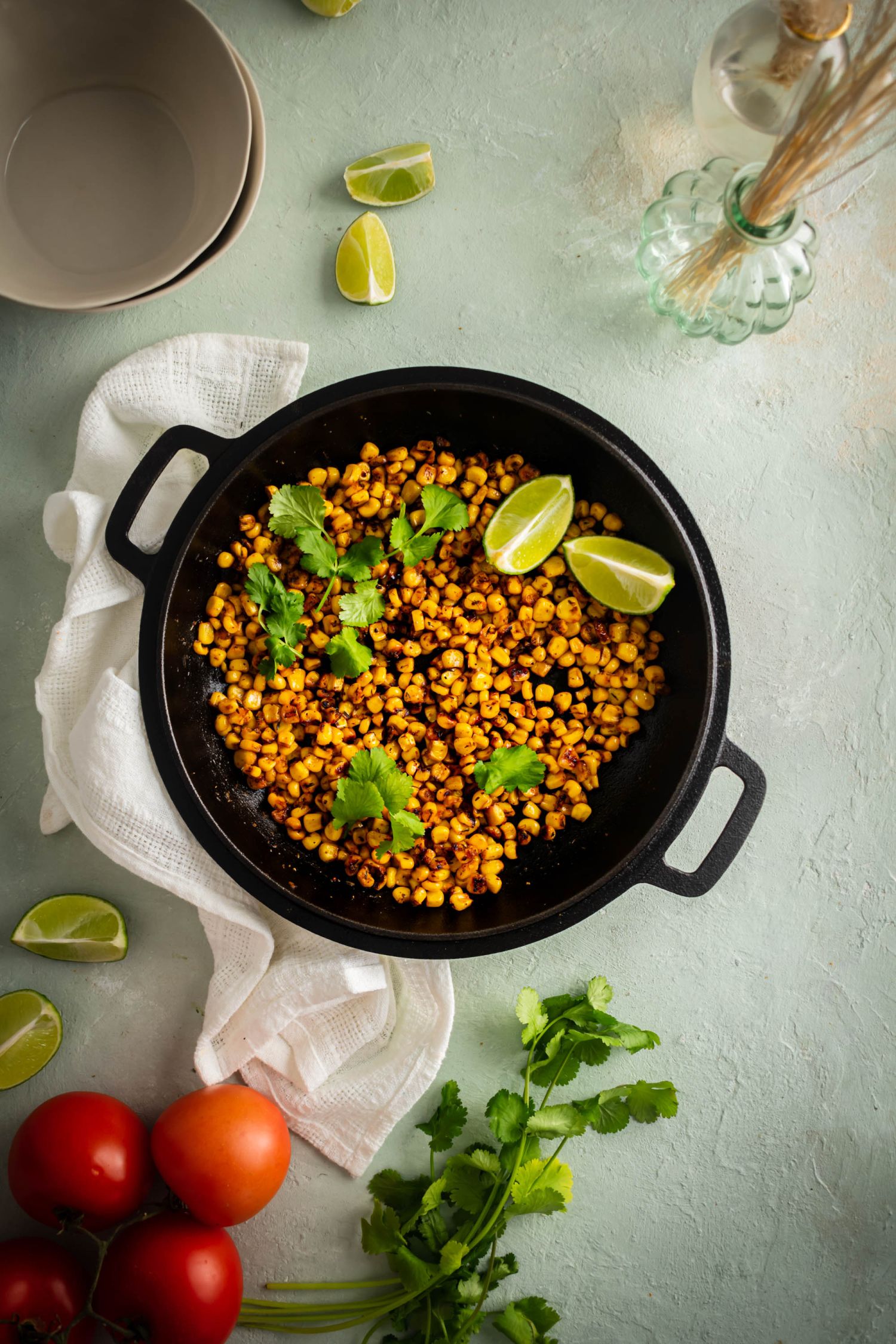 Cast iron skillet with blackened corn cooked in butter and spices with tomatoes, limes, and cilantro on the side.