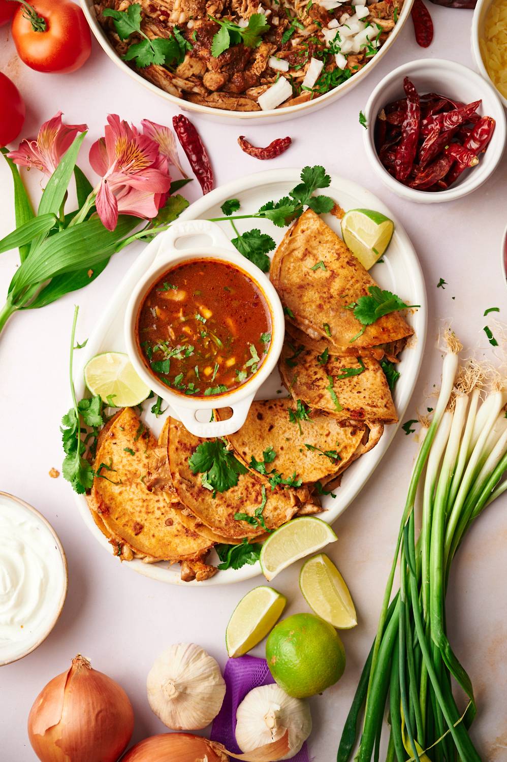 Chicken birria tacos with consome served on a plate with limes, cilantro, and dried chile peppers.