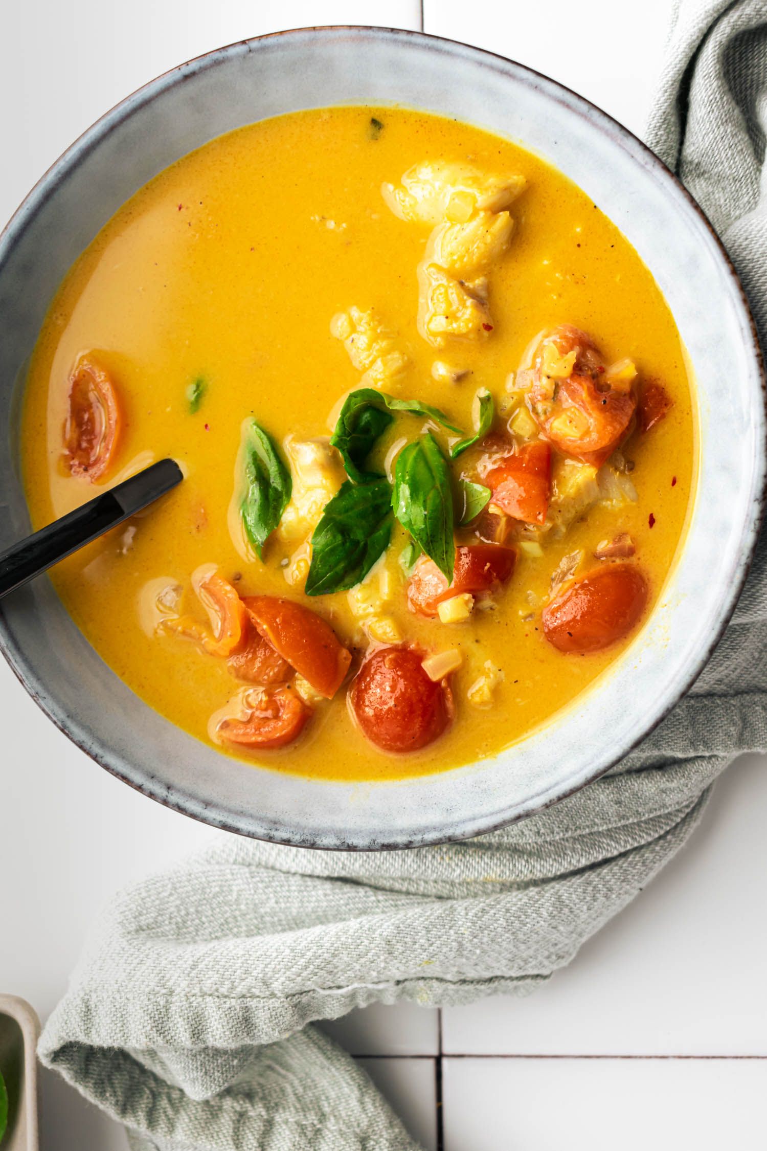 Bowl of fish curry with fresh herbs, cherry tomatoes, and a creamy curry coconut broth with a spoon.