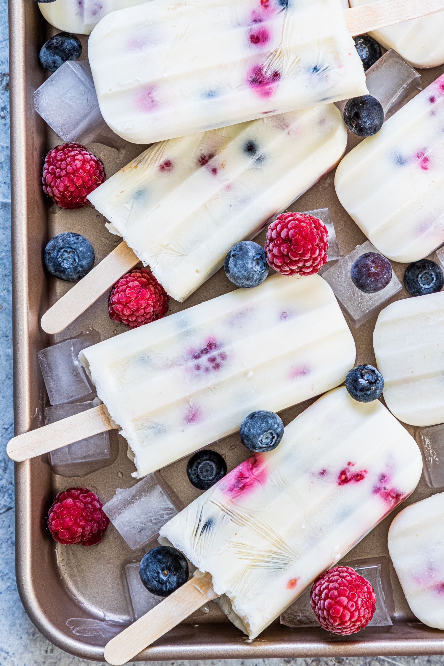 Creamy greek yogurt and fruit popsicles on a tray with frozen raspberries and blueberries.