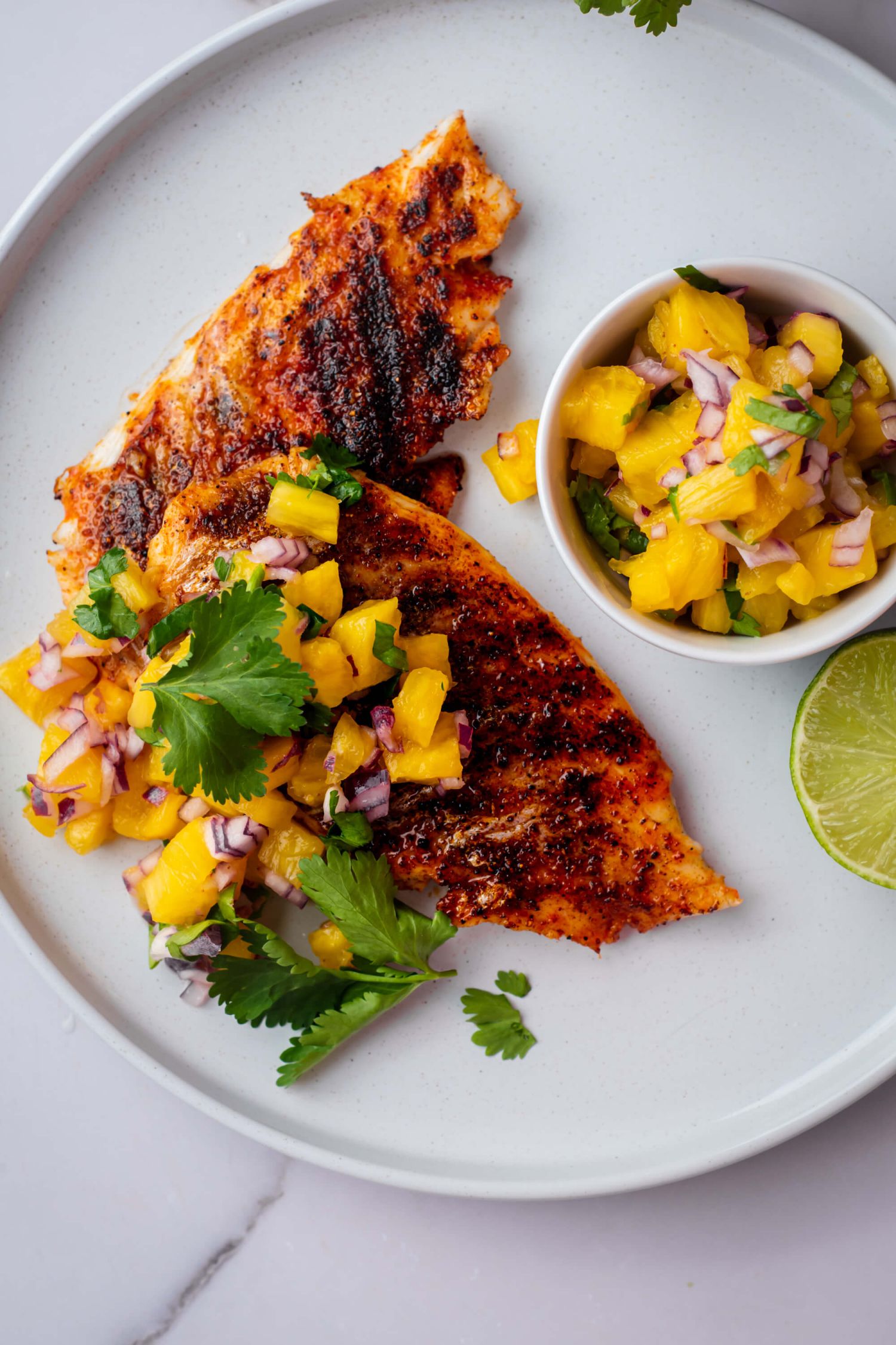 Two grilled mahi mahi fillets with a dark red spice rub and fresh pineapple salsa on a white plate.