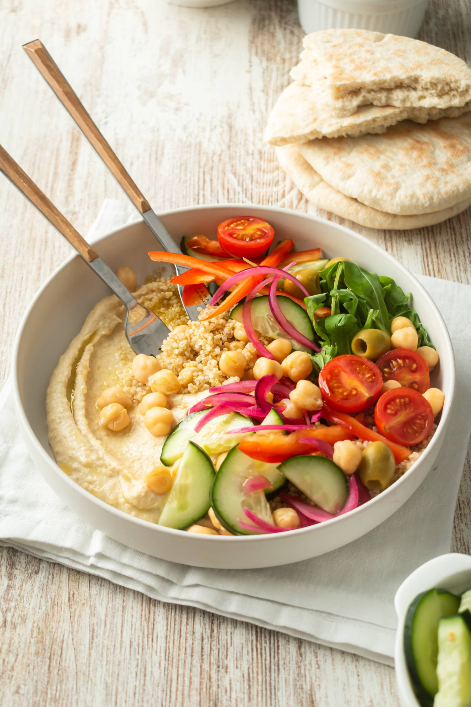 Hummus quinoa bowl with raw vegetables and olives in a white bowl with pita bread.