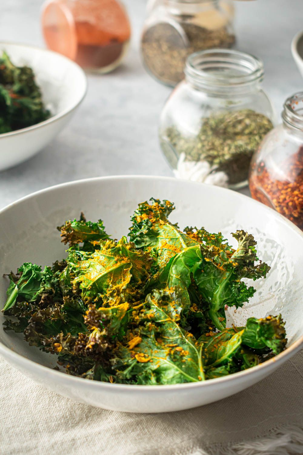 Crispy baked kale chips in a bowl with nutritional yeast on top and spices in the background.