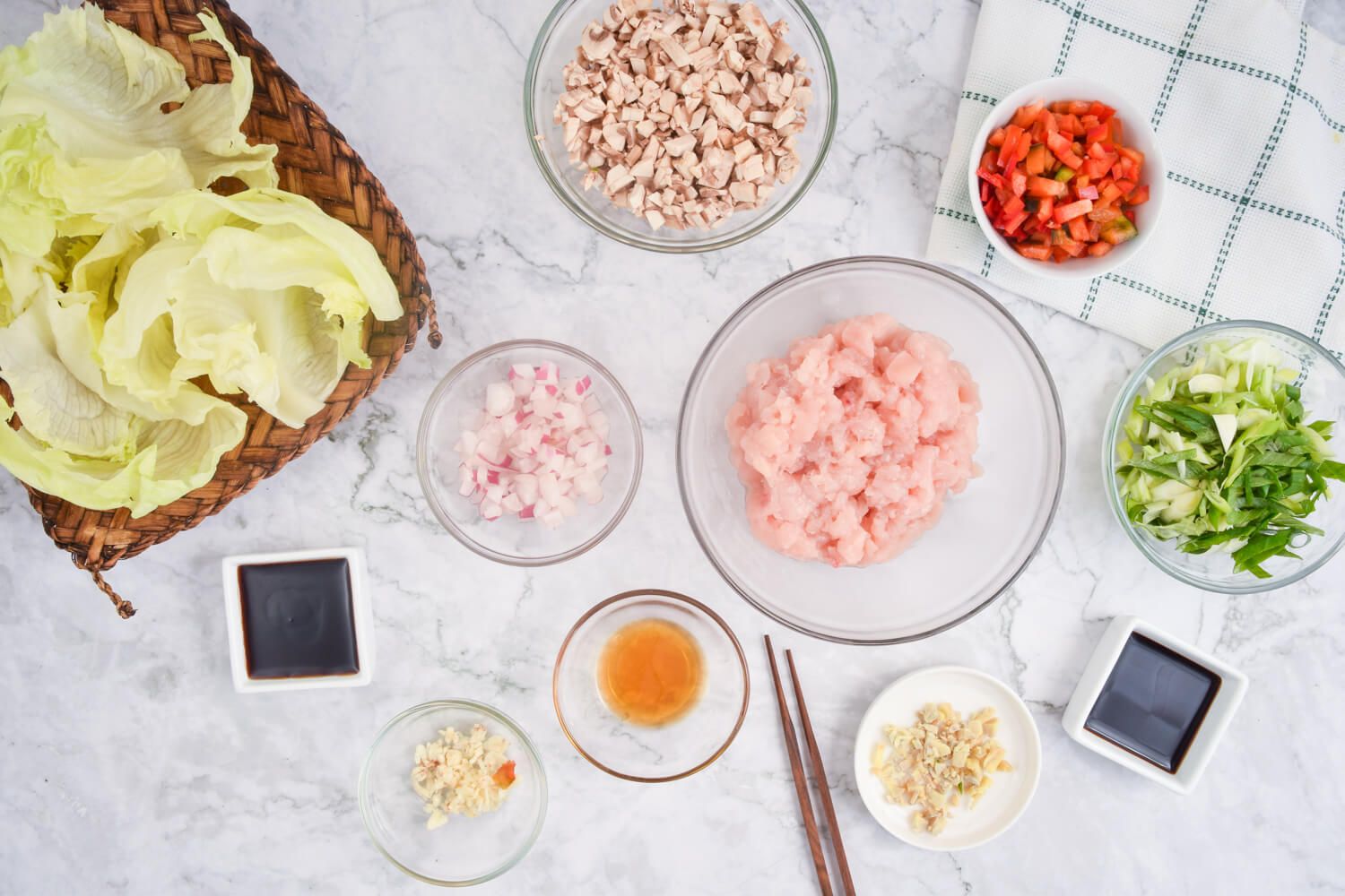 Ingredients for chicken lettuce wraps on a marble background including chicken, lettuce, mushrooms, and red peppers.