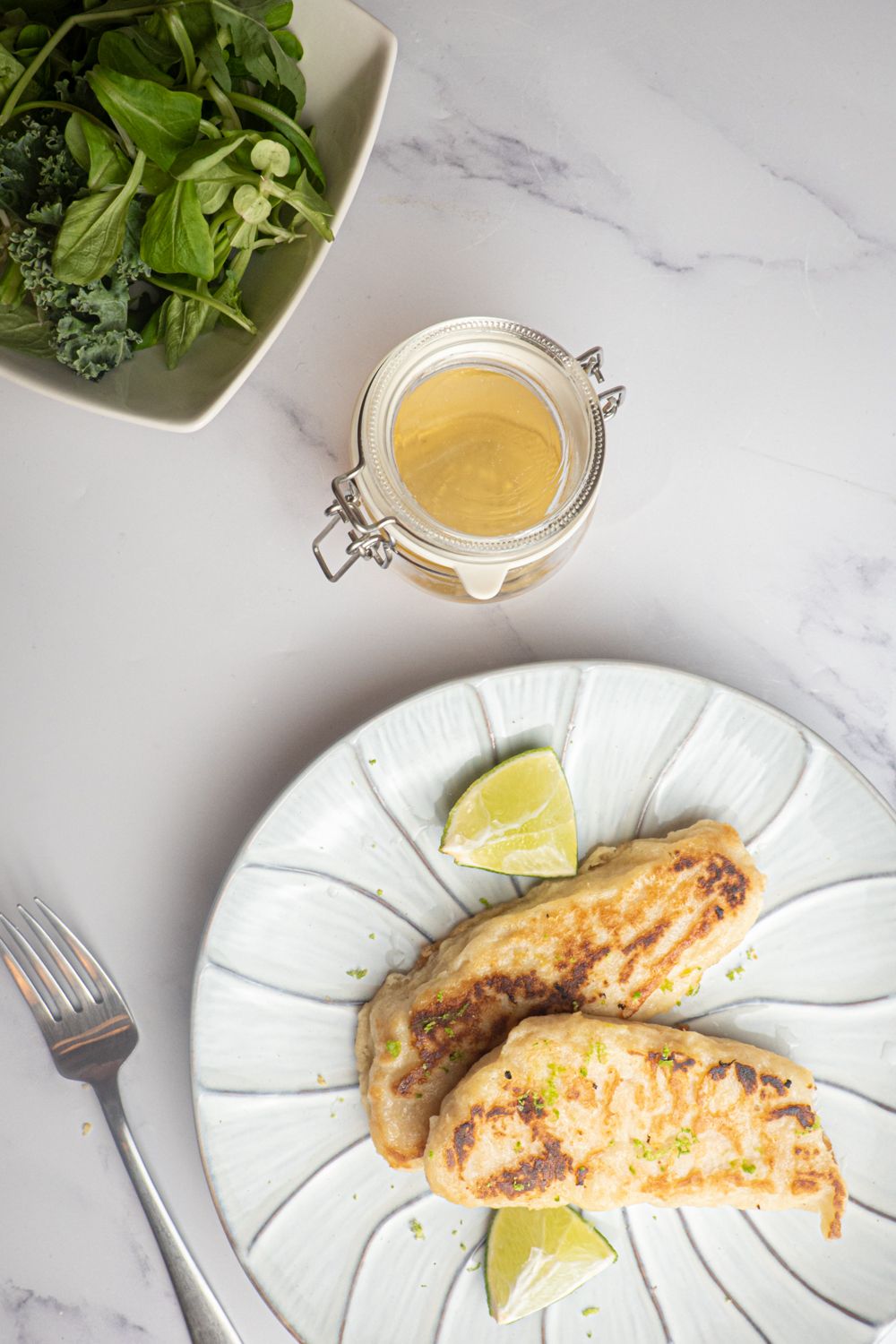 Tilapia with honey and lime on a plate with a bowl of greens and a fork.