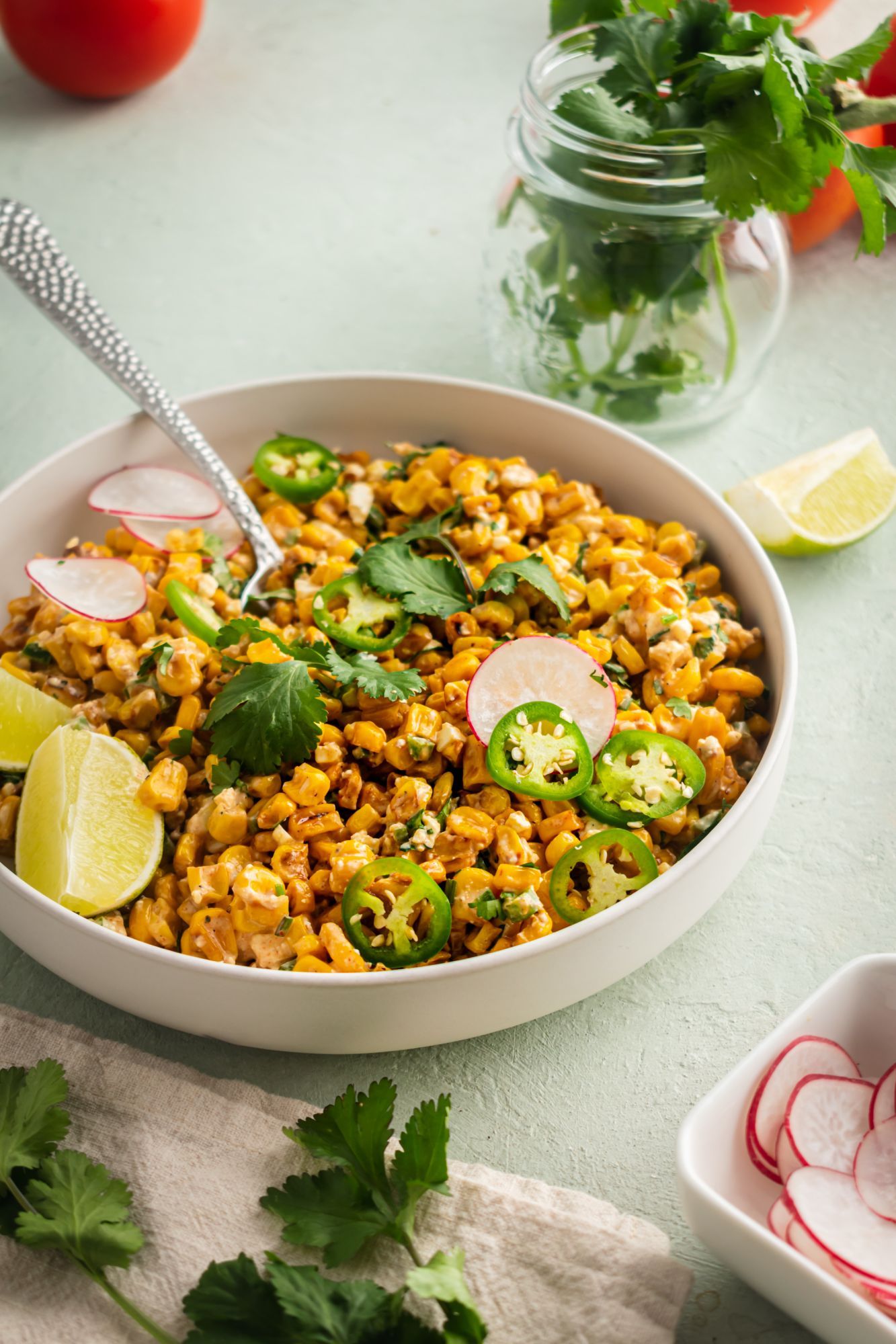 Mexican corn salad with a creamy Cotija cheese and lime dressing with sliced jalapenos, cilantro, radish, and lime in a bowl.