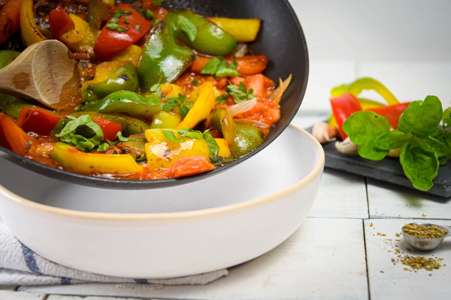 Italian pepperonata in a skillet with peppers, tomatoes, and onions being added to a bowl with a wooden spoon.