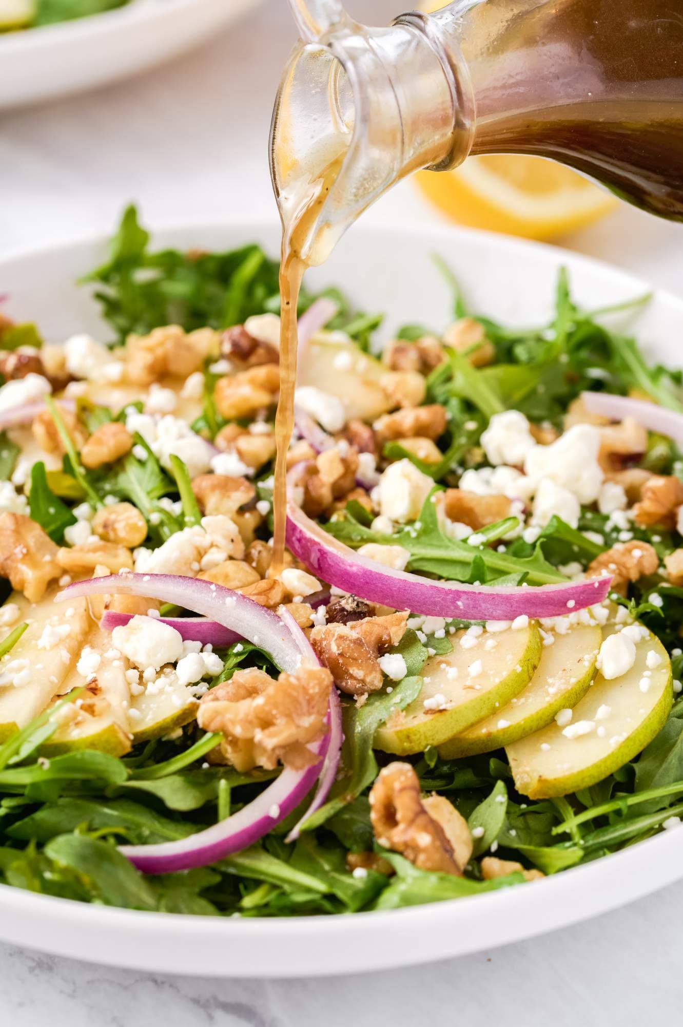 Arugula goat cheese salad with sliced pears, red onions, and walnuts with vinaigrette being poured on top.