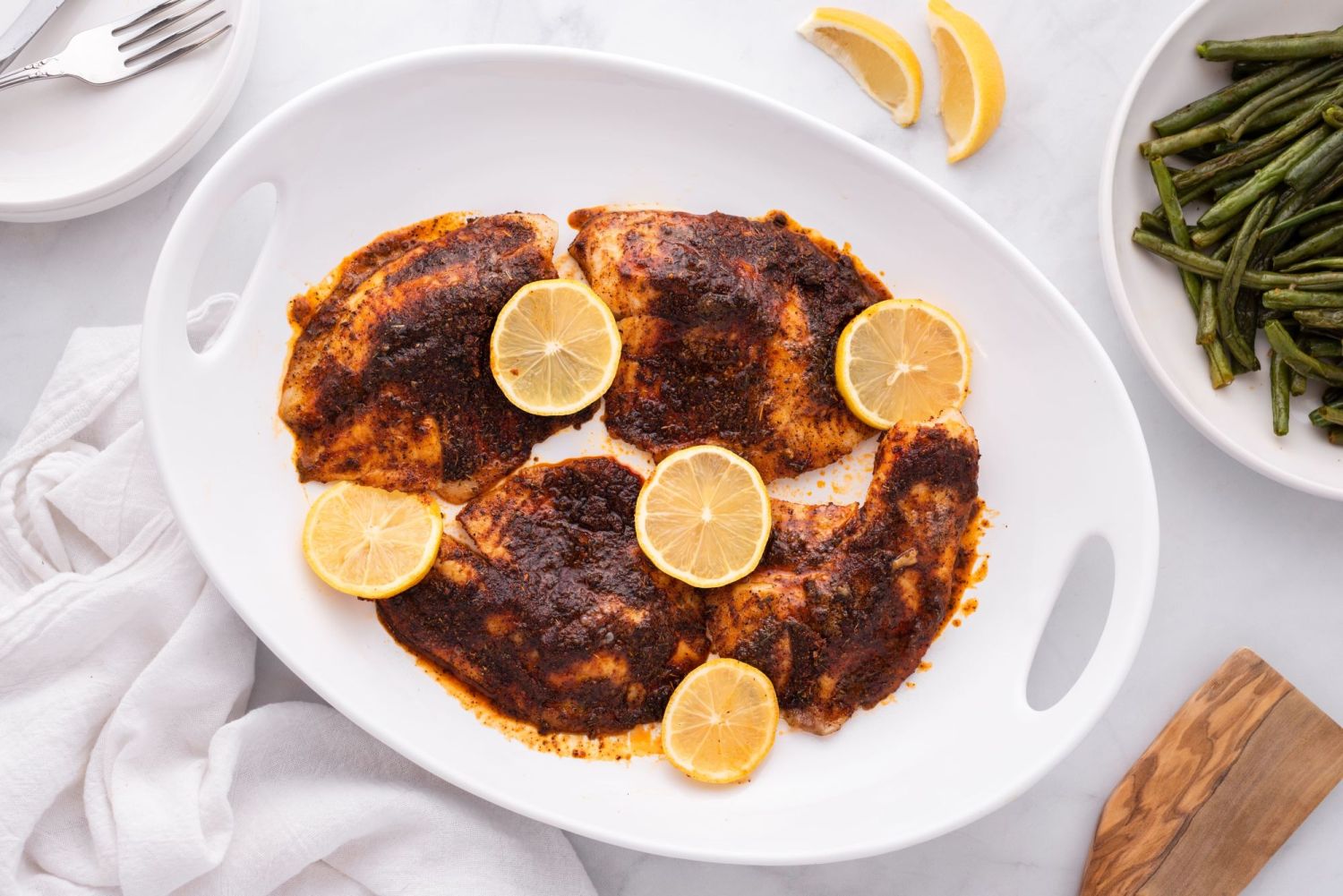 Blackened tilapia filets served on a white plate with lemon slices and green beans.