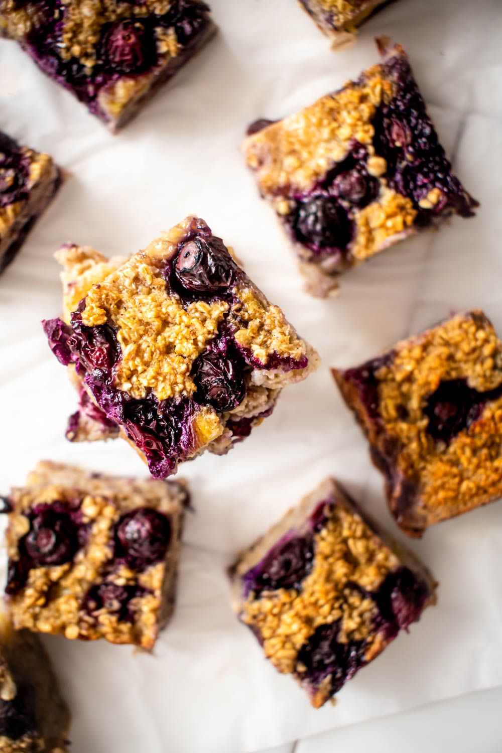 Baked blueberry banana oatmeal cut into squares with rolled oats and blueberries on parchment paper.