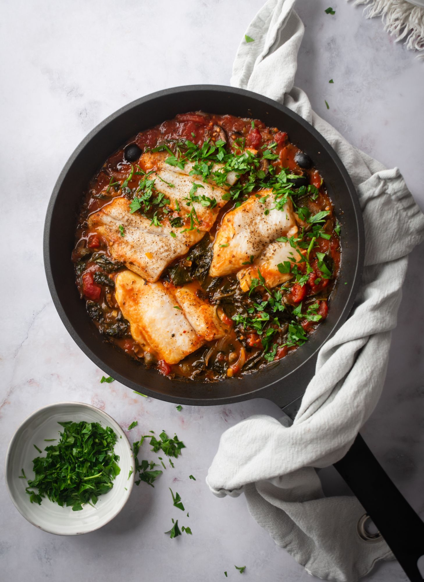 Mediterranean baked in a skillet with tomatoes, spinach, black olives, capers, and parsley.