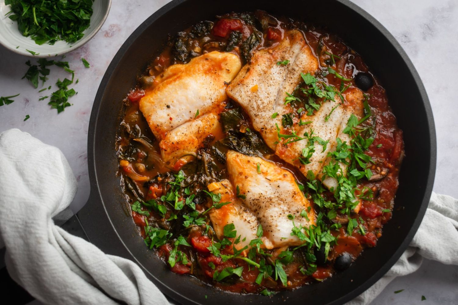 Cast iron skillet with Mediterranean white fish with tomatoes, olives, capers, garlic, spinach, and fresh parsley.