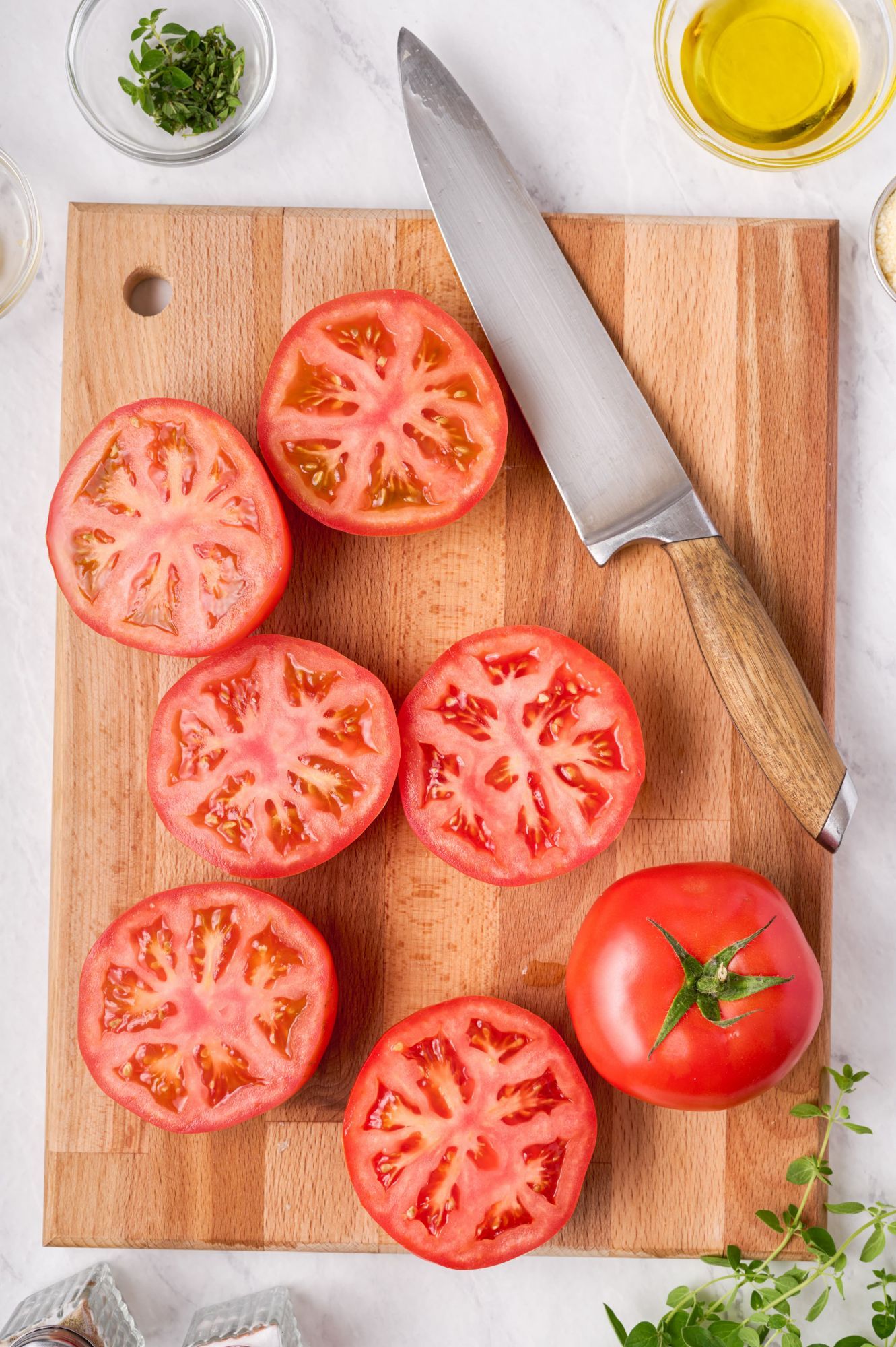 Sliced tomatoes on a cutting board with herbs, Parmesan cheese, and olive oil on the side.