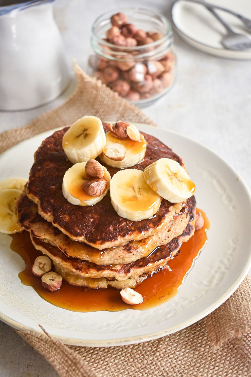 Oat pancakes stacked on a plate with sliced bananas, hazelnuts, and maple syrup.