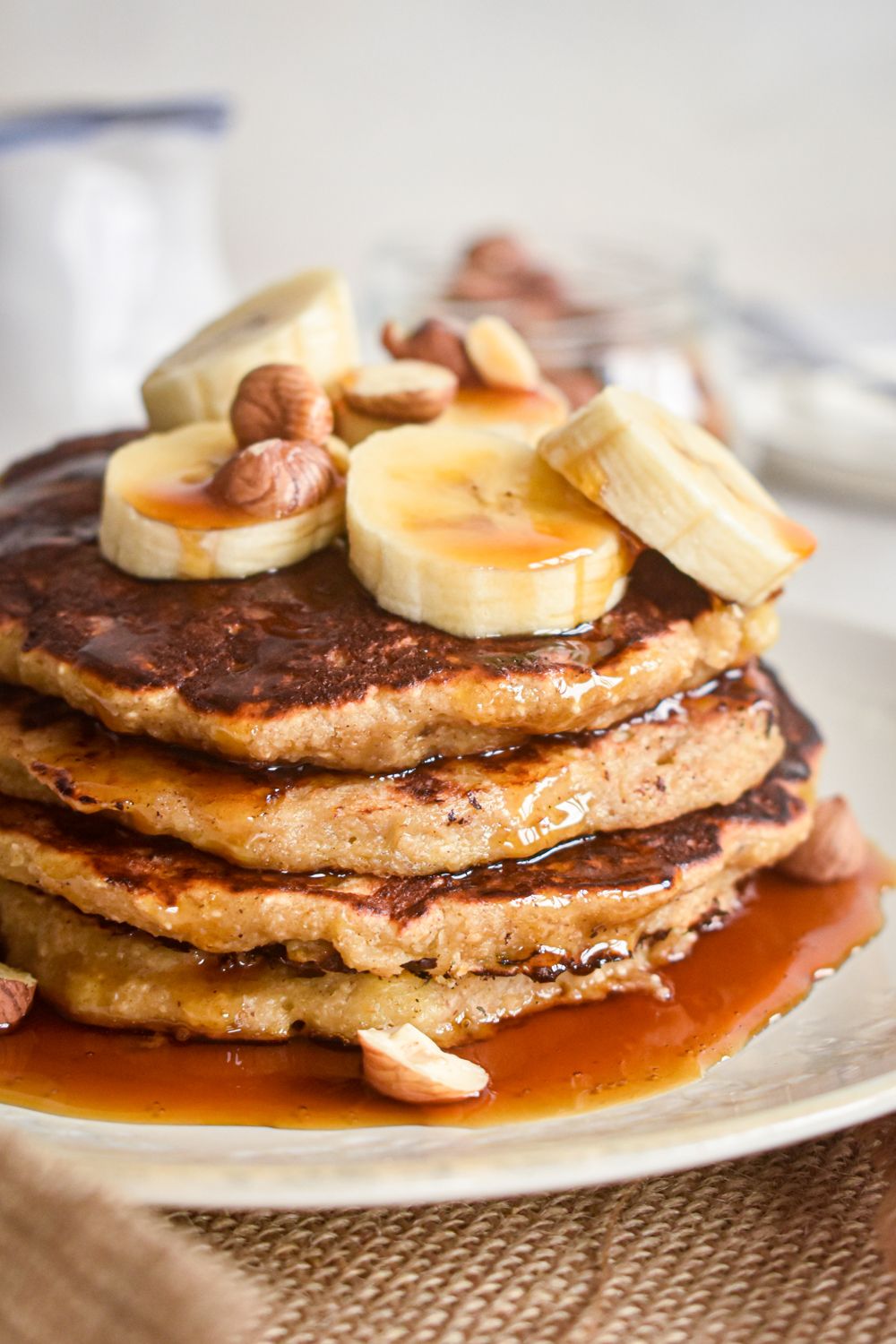 Banana oatmeal pancakes on a plate with sliced bananas, maple syrup, and chopped hazelnuts. 