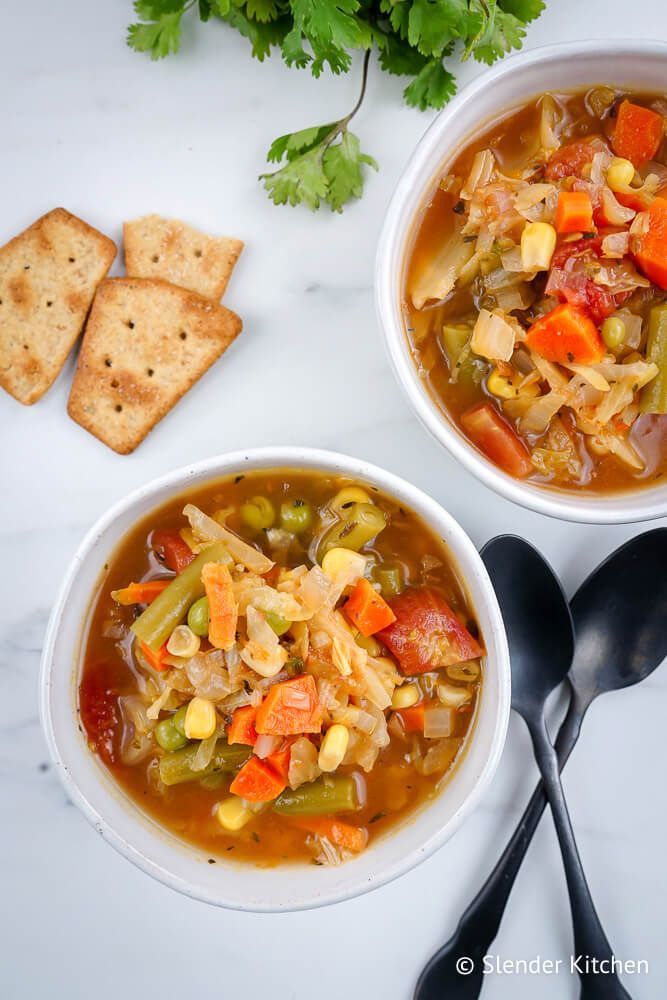 Homemade vegetable soup with cabbage, carrots, tomatoes, corn, and peas in two bowls with spoons.