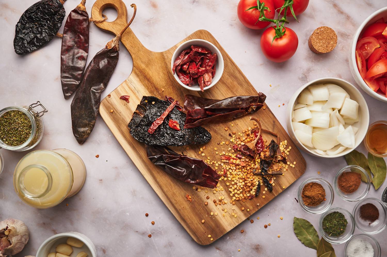 Dried chilies including guajillo, arbol, and ancho peppers on a cutting board with onions, spices, and tomatoes.