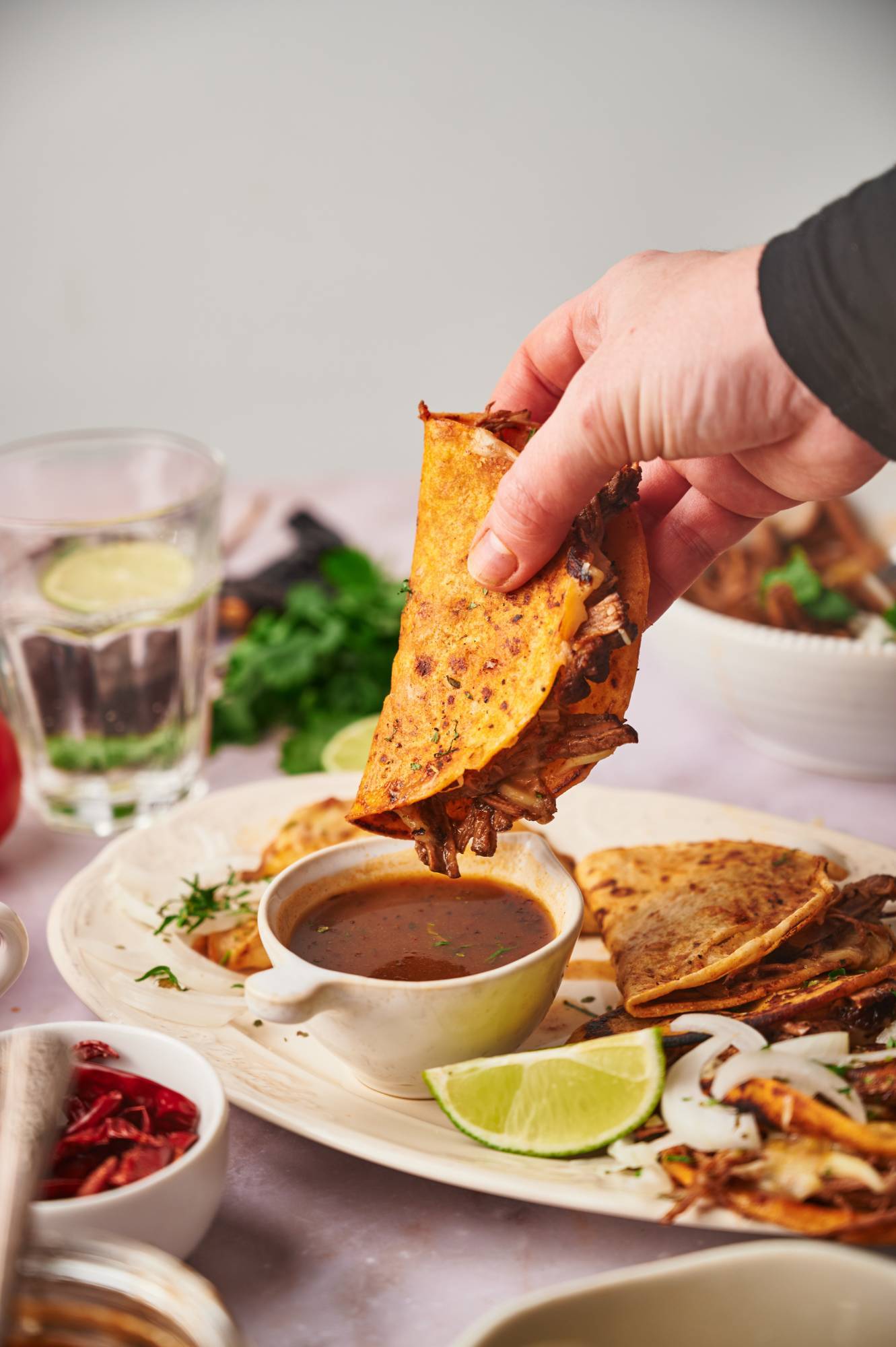 Beef birria taco being dipped into a cup of consome with lime, white onion, and cilantro.