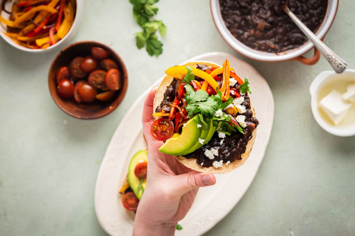 Baked bean tostada with salsa black beans, fajita vegetables, queso fresco, and avocado being held up by a hand.