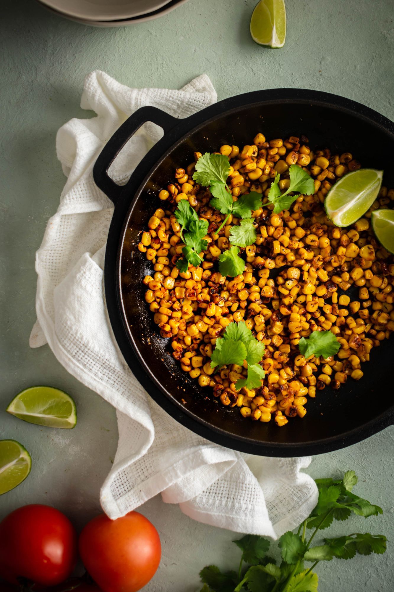 Corn with blackening seasoning in a cast iron skillet with cilantro, limes, and tomatoes on the side.