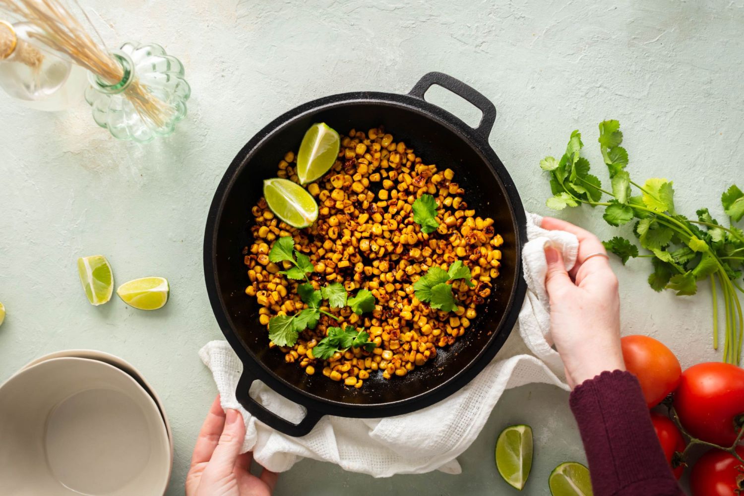 Charred blackened corn with lime wedges and cilantro leaves in a black cast iron skillet being held by two hands,