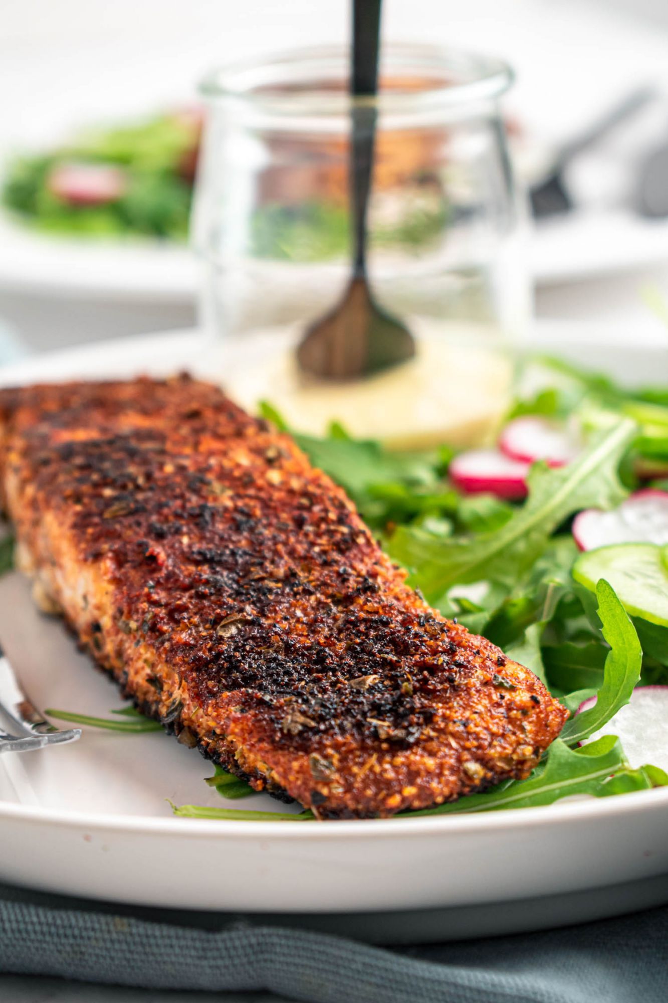 Salmon cooked in blackening seasoning on a plate with greens, cucumbers, and radishes.