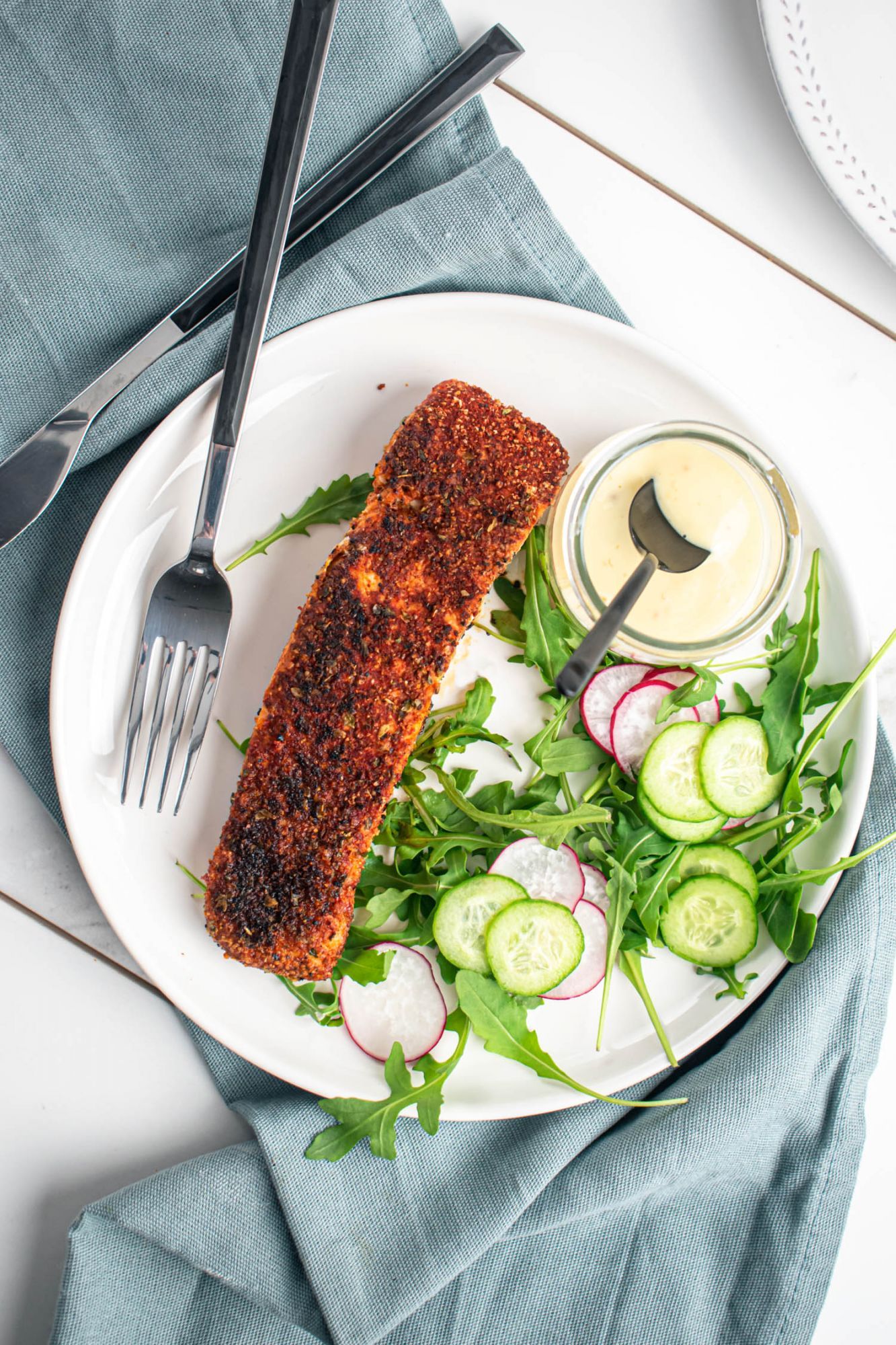 Cast iron cooked blackened salmon on a plate with jar of aioli, cucumbers, and salad greens.