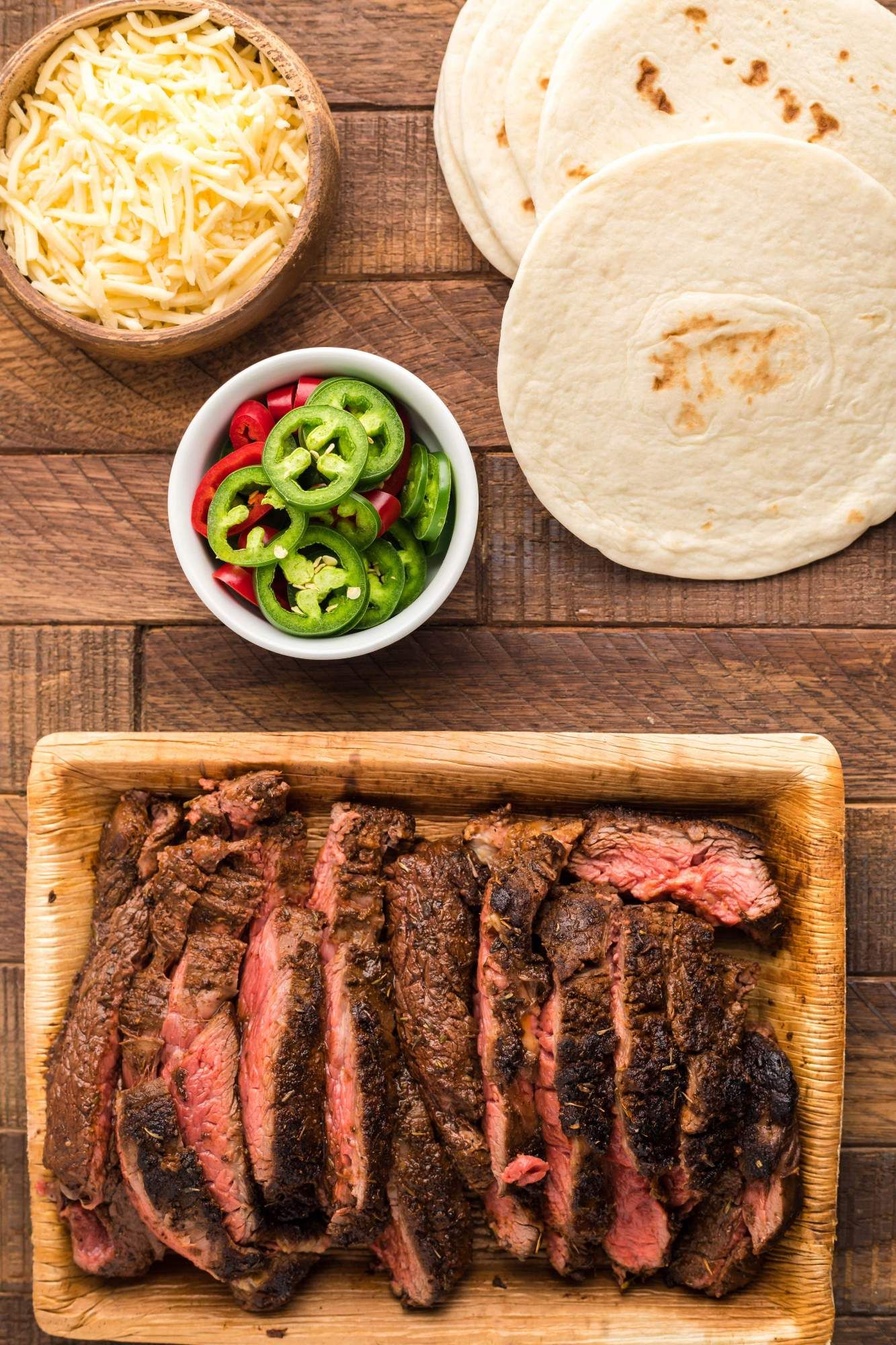 Sliced blackened steak with jalapenos, flour tortillas, and shredded cheese on the side.