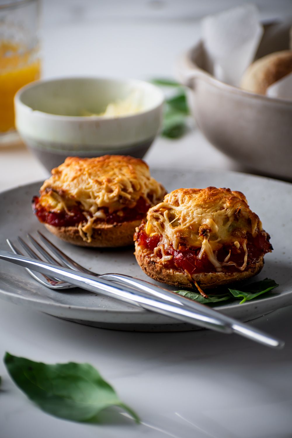 Two English muffins topped with scrambled eggs, tomato sauce, and melted cheese for breakfast pizzas on a plate with basil.