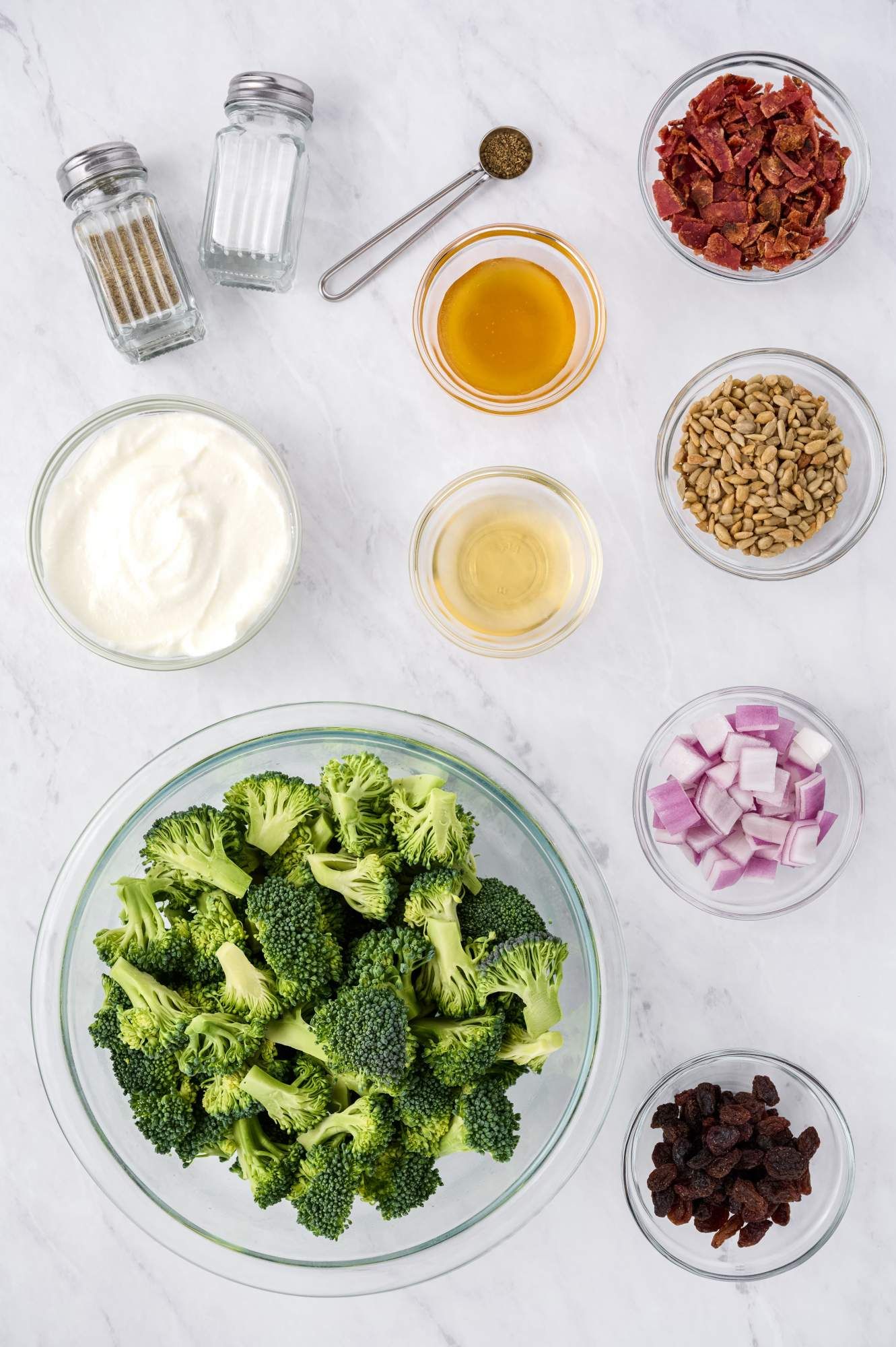 Ingredients for broccoli salad including fresh broccoli, yogurt, mustard, bacon, onions, raisins, and sunflower seeds.