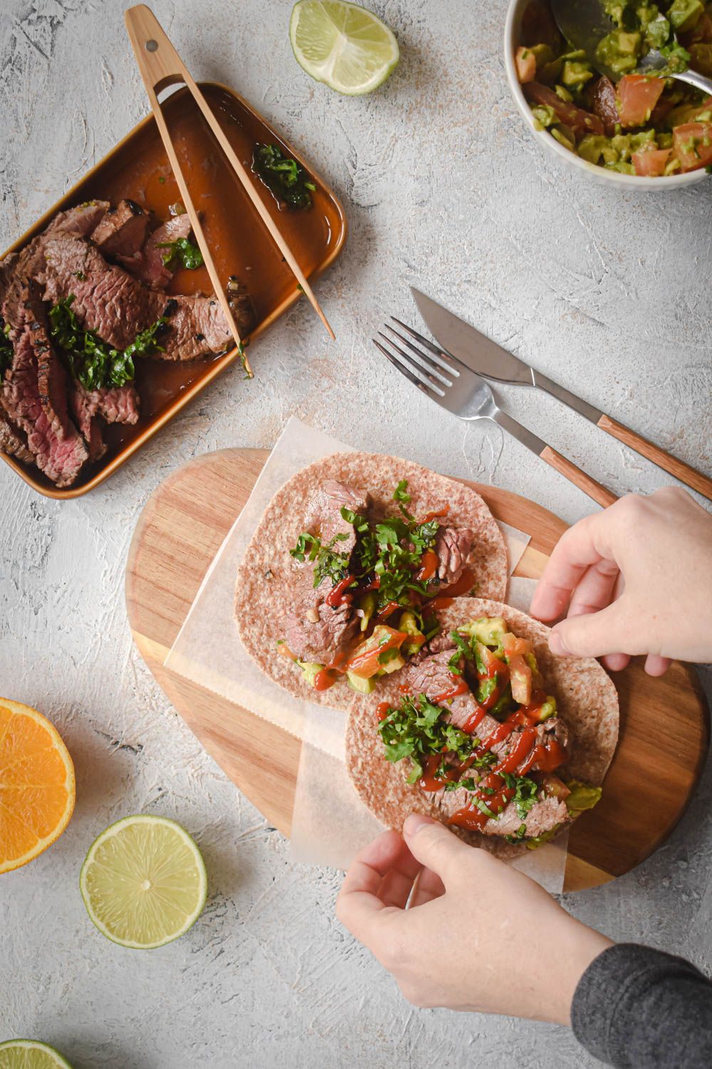 Grilled carne asada served in tacos with guacamole, cilantro, and salsa being picked up by two hands.