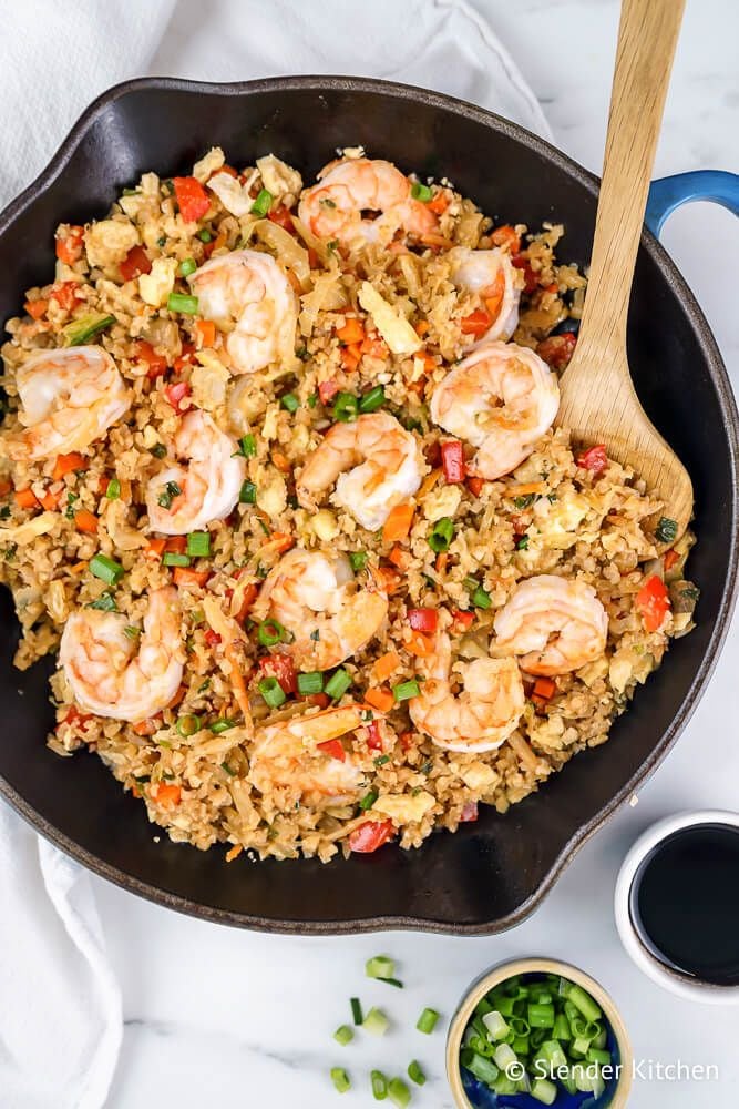 Shrimp cauliflower fried rice in a skillet with vegetables, a wooden spoon, and green onions.