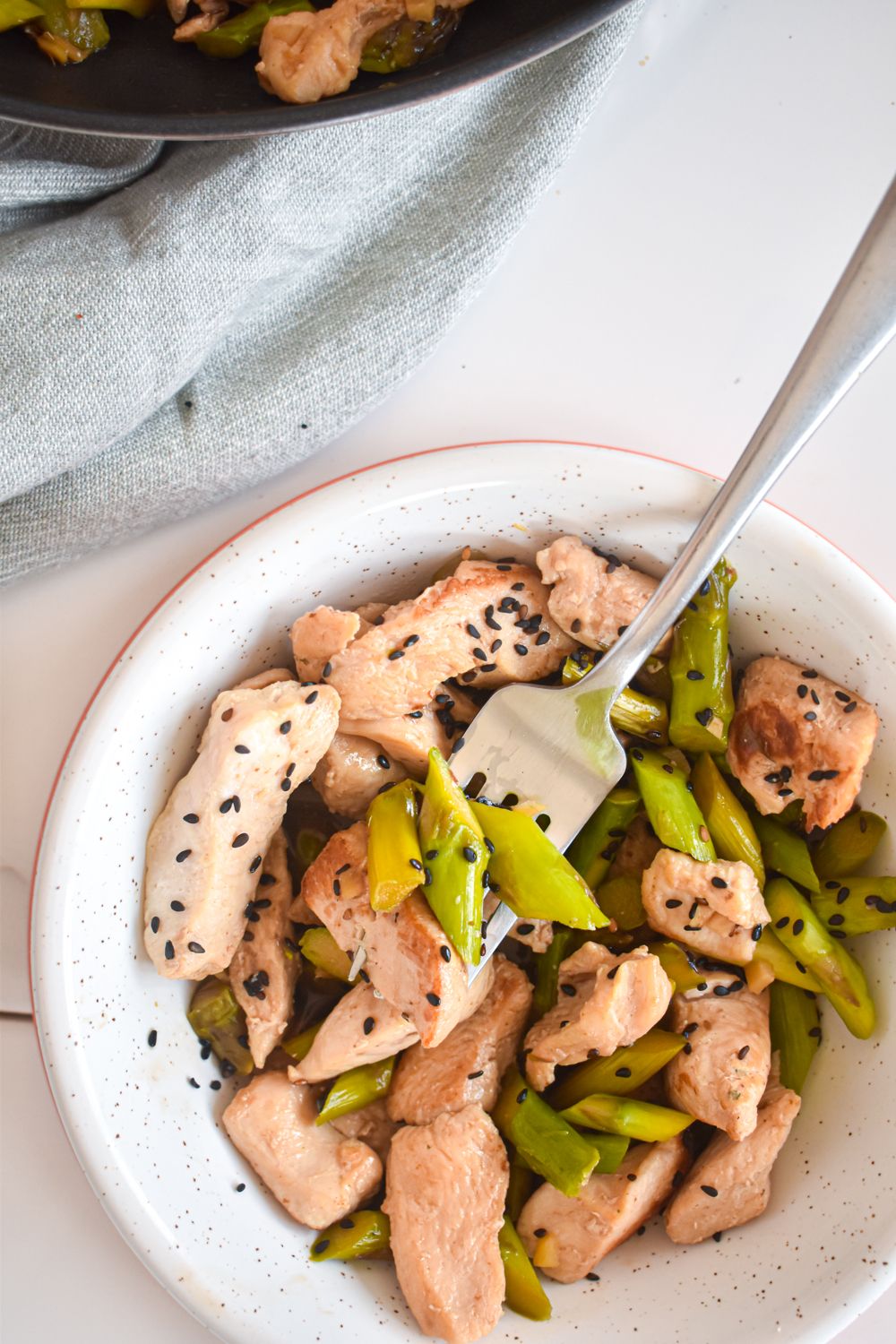 Asparagus and chicken stir fry with sesame seeds in a bowl with a skillet on the side.