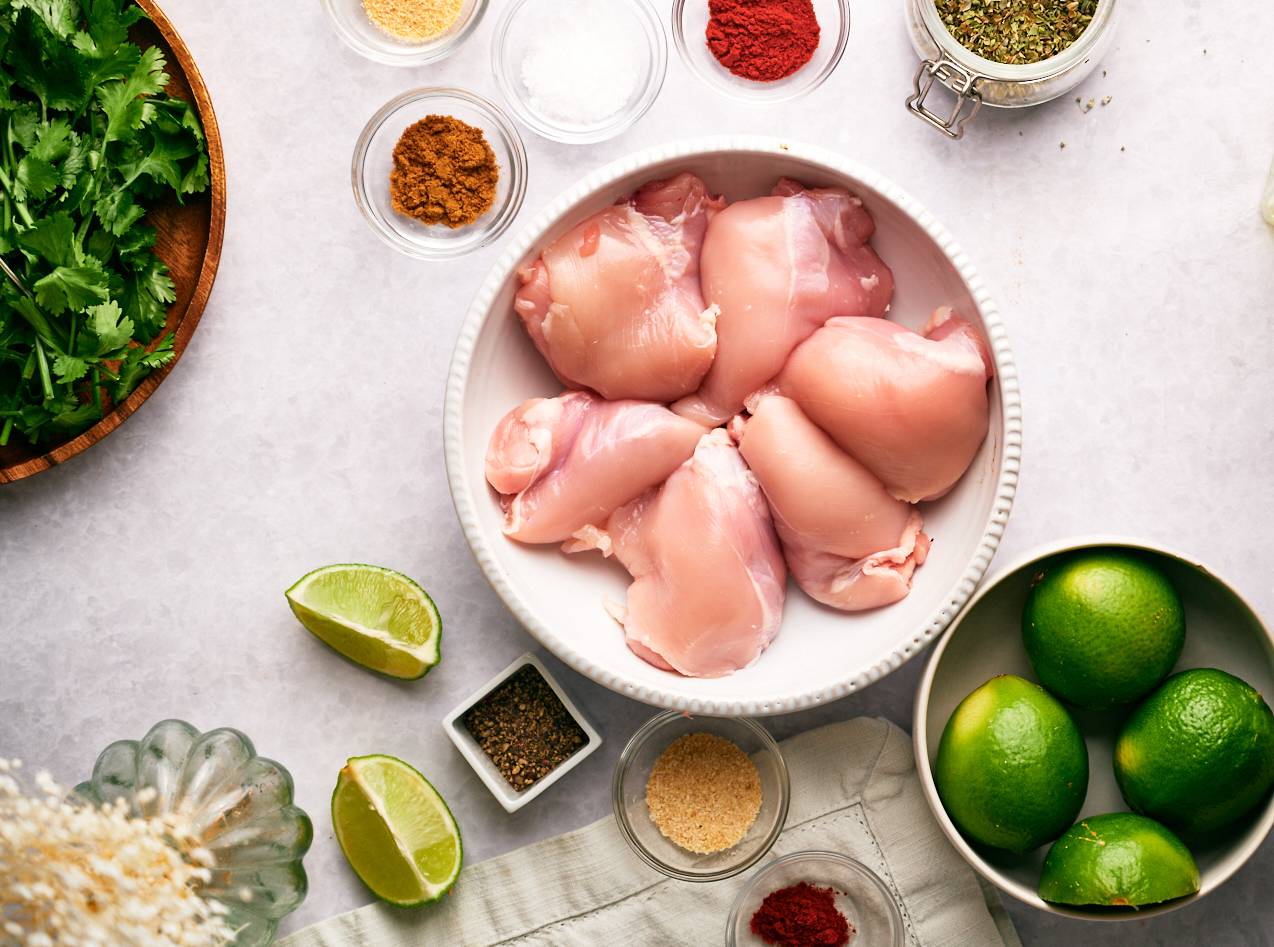 Ingredients for chicken carnitas including chicken thighs, cilantro, spices, and limes.