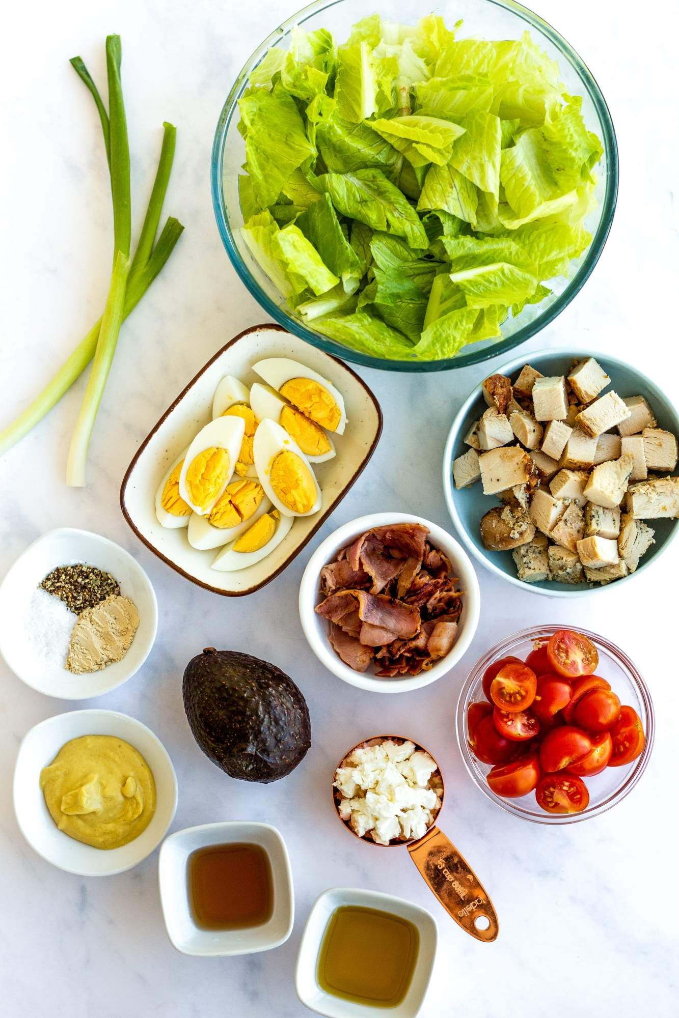 Ingredients for a Chicken Cobb Salad including chopped lettuce, eggs, bacon, chicken, tomatoes, and avocado.