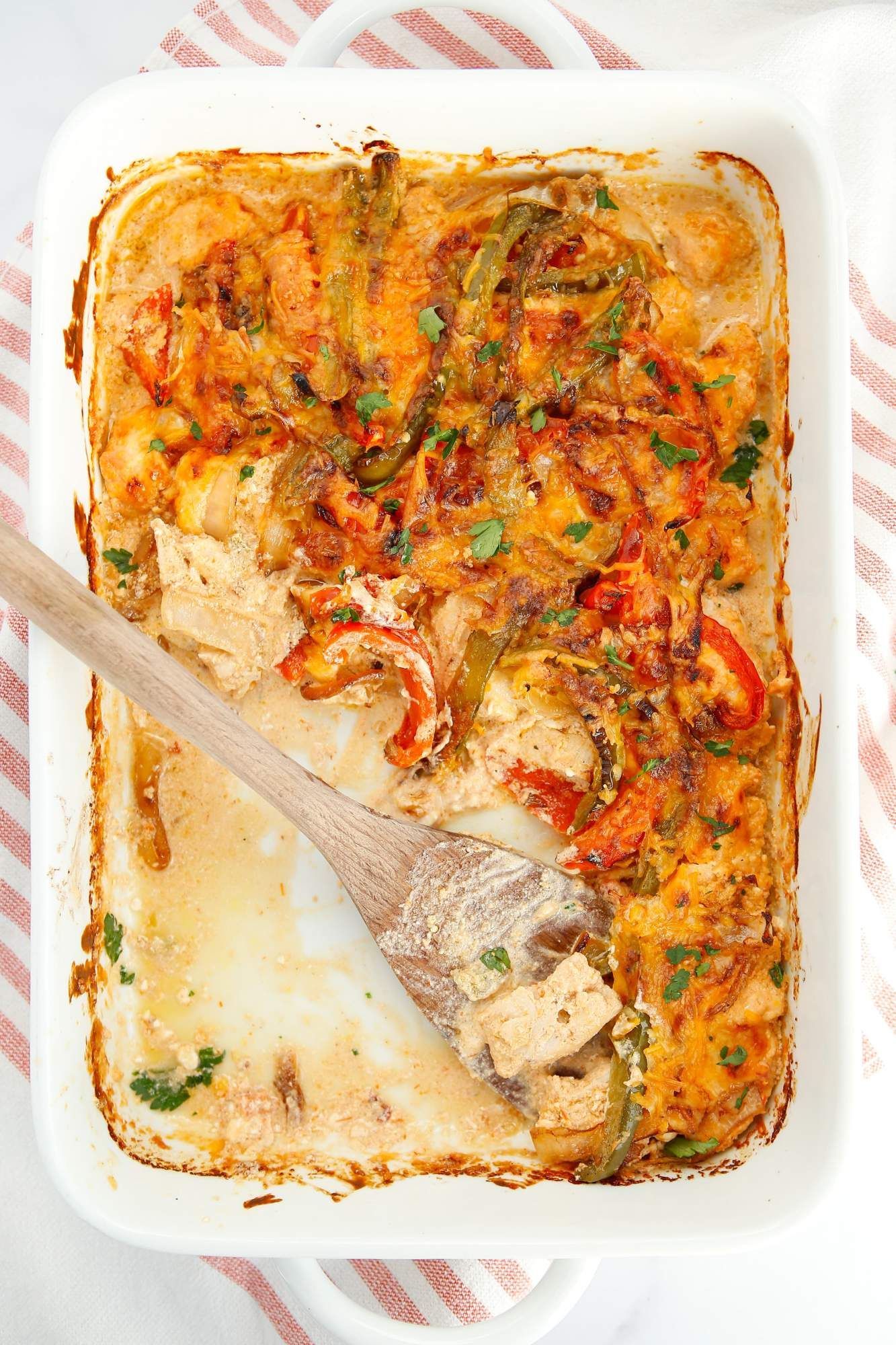 Chicken fajita bake with peppers, onions, chicken breast, and a creamy sauce in a casserole dish with a wooden spoon.