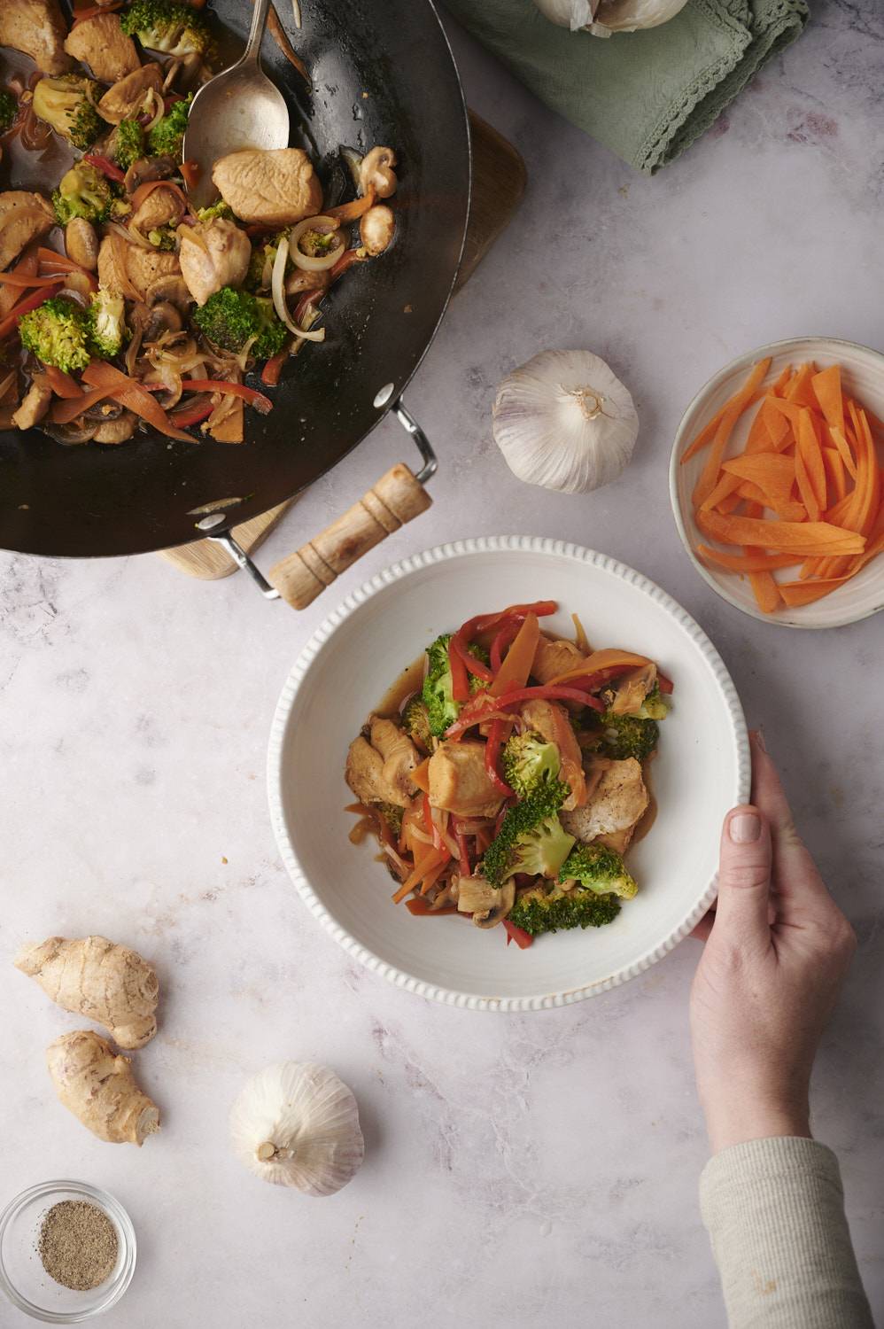 Stir fried chicken and vegetables in a white bowl and in a wok with fresh carrots on the side.