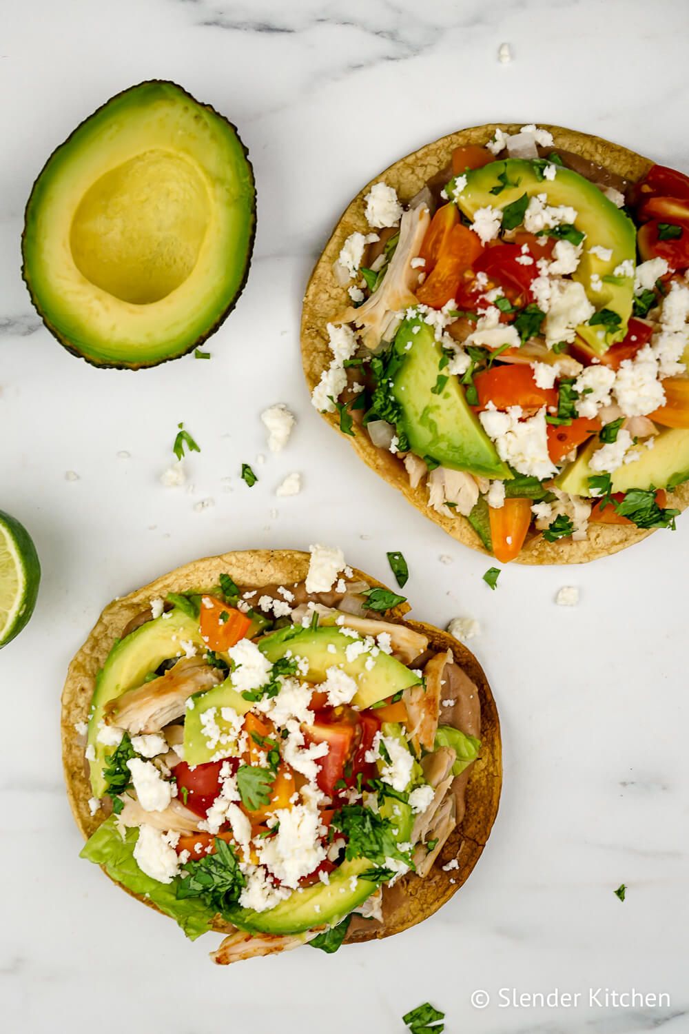 Tostada with chicken, lettuce, tomatoes, and avocado on a marble board.