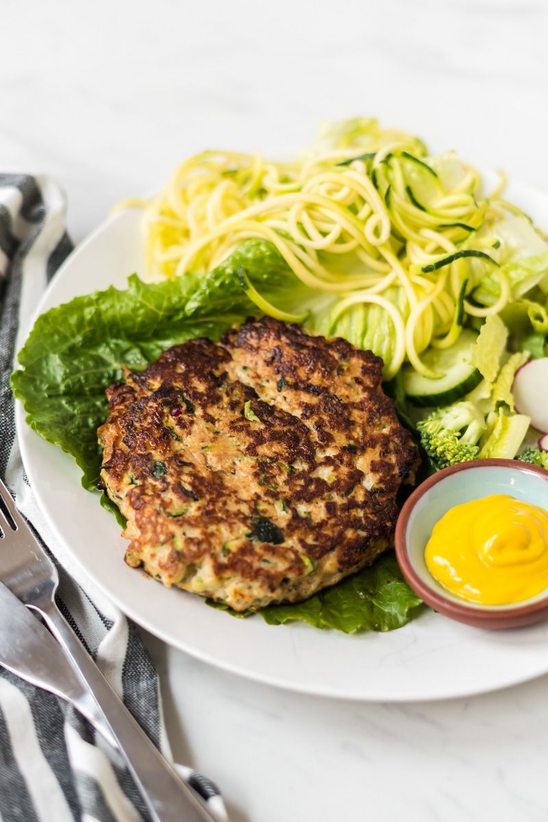 Shredded zucchini and chicken burgers on a plate with greens and zucchini noodles.