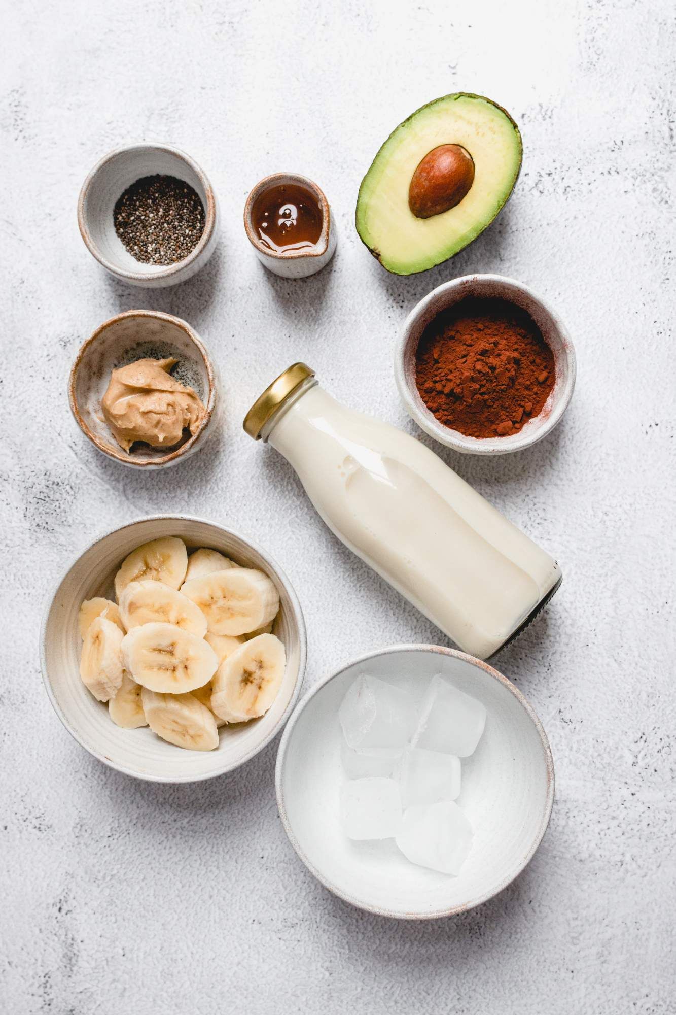 Ingredients to make an avocado smoothie including avocado, banana, cocoa powder, almond butter, and almond milk.