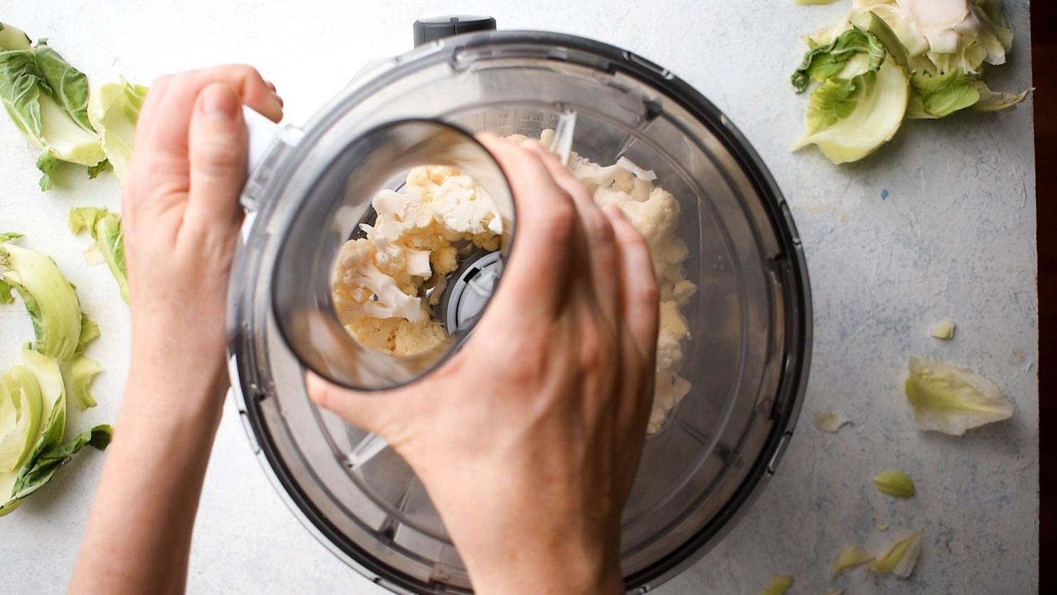 Cauliflower florets being added to a food processor to make cauliflower rice with the outer leaves on the side.