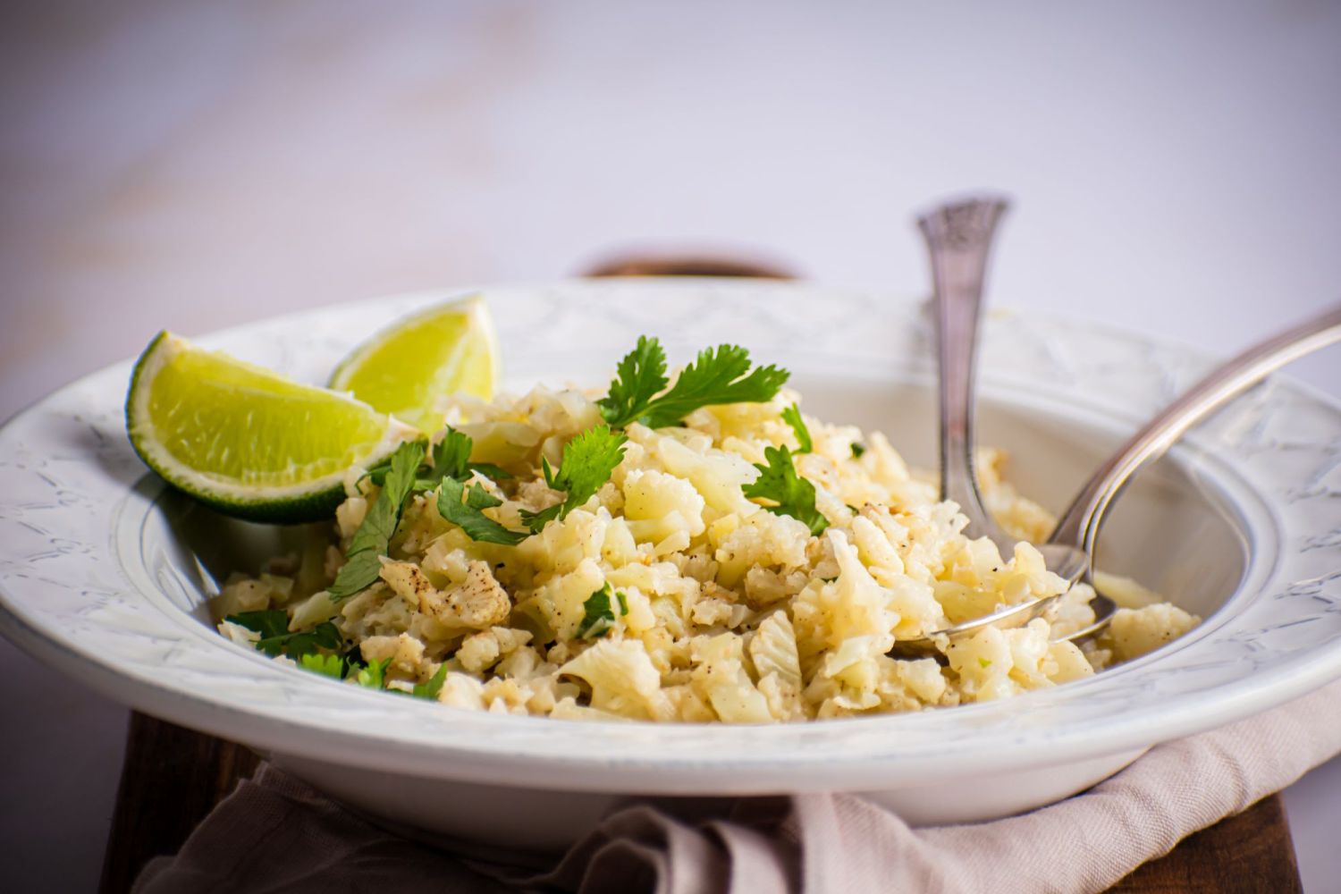 Coconut riced cauliflower with cilantro leaves and fresh lime in a bowl on a cutting board.