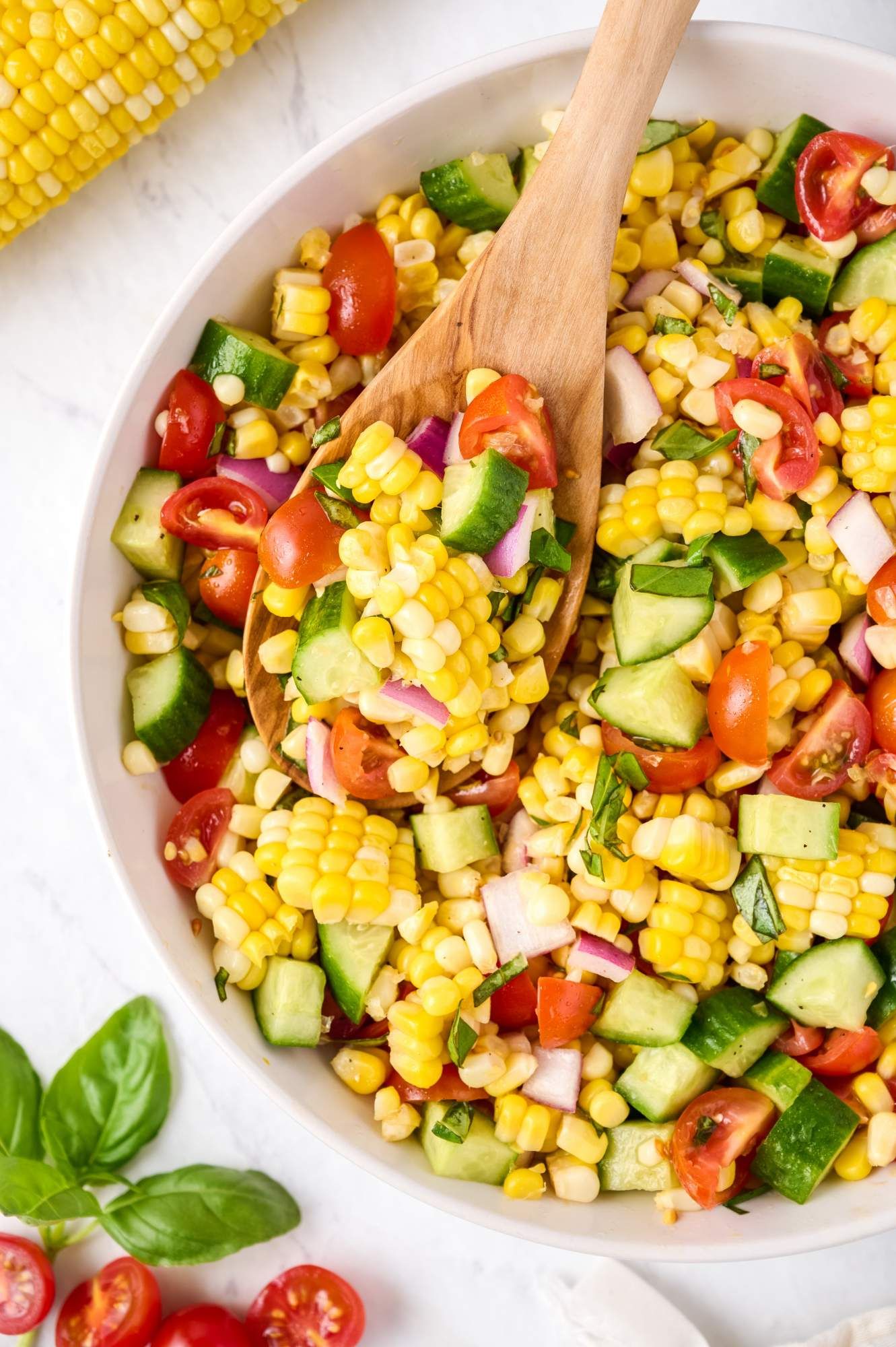 Fresh corn salad with tomatoes, cucumbers, basil, and dressing in a large white bowl with a wooden spoon.