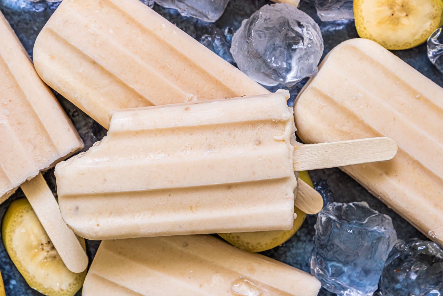 Banana ice pops with yogurt and maple syrup stacked on a tray with ice cubes.